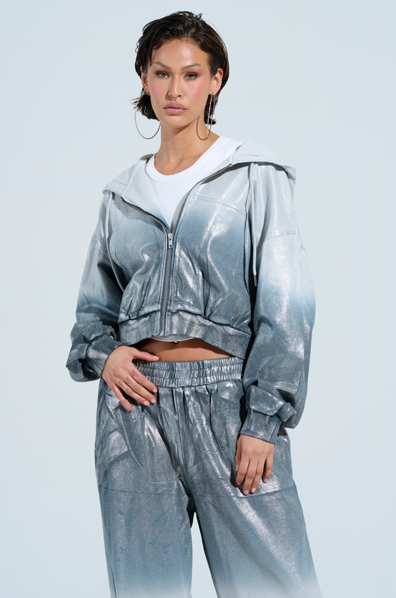 Wearing the SHINING DOWN ON YOU SWEATSHIRT, a person stands against a light blue background, showcasing an eye-catching, shiny silver-blue gradient tracksuit paired with a white T-shirt. The loungewear ensemble has an oversized fit, including a zip-up hoodie and loose-fitting pants that add flair to their style. Their look is further enhanced by hoop earrings and short hair.