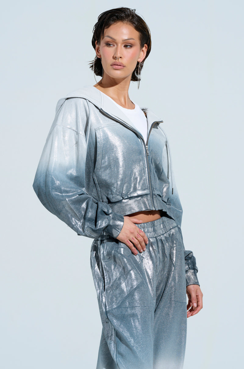 Against a light background, an individual confidently poses in an oversized tracksuit with a metallic finish, featuring the SHINING DOWN ON YOU SWEATSHIRT in a shiny silver gradient. Complementing the look is a white shirt underneath. Their short, styled hair adds to their side-glancing stance.