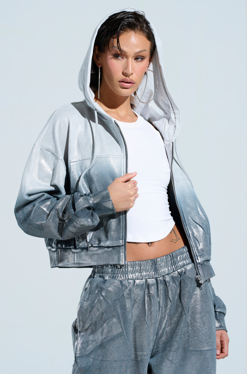 A person wearing the SHINING DOWN ON YOU SWEATSHIRT, characterized by its metallic silver finish and oversized fit, along with matching pants, stands confidently against a light blue background. They are styled with a white crop top and large hoop earrings.