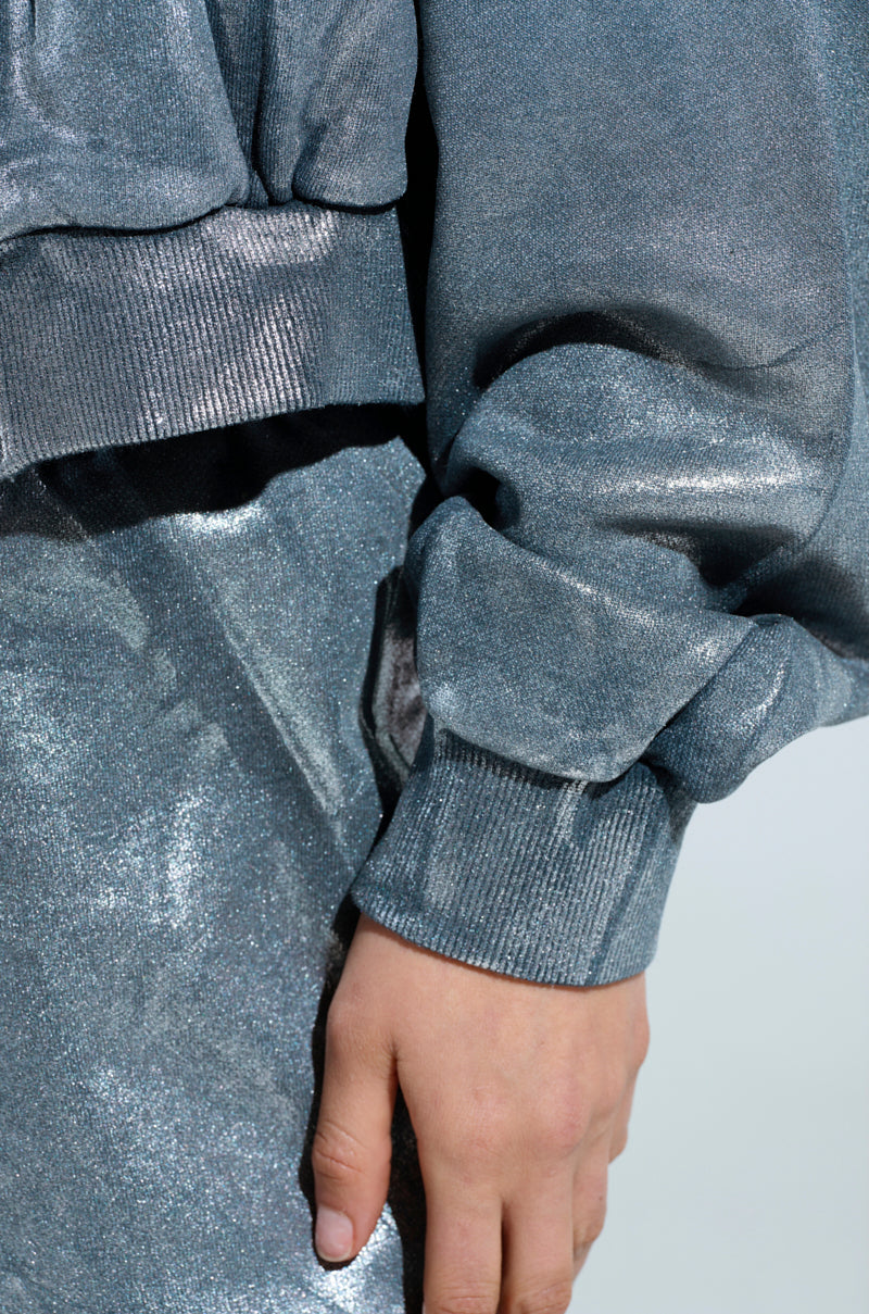 Close-up of a person wearing the SHINING DOWN ON YOU SWEATSHIRT, an oversized metallic silver outfit with ribbed cuffs on the sleeves. The hand rests against the hip, accentuating the fabric's texture and shimmer under the light, seamlessly merging loungewear comfort with a stylish metallic sheen.
