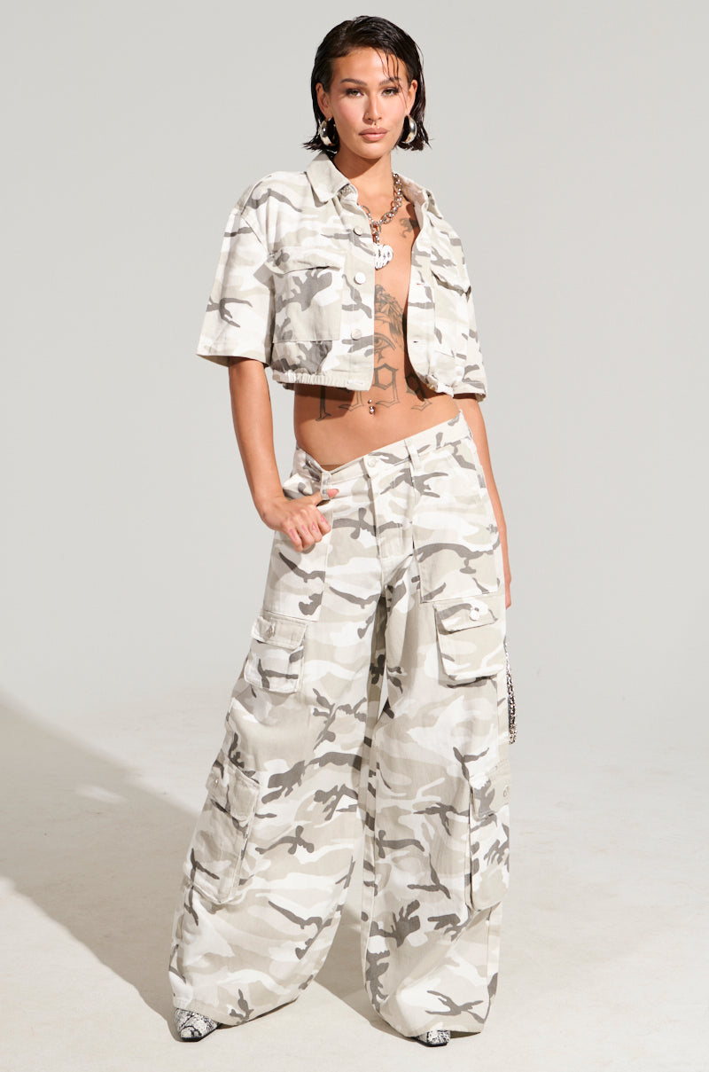 A person stands confidently in a light camouflage outfit, showcasing a cropped jacket paired with YOUR UP NEXT CAMO CARGO PANT featuring cargo pockets and a high-rise fit. They have short hair, visible tattoos, and wear a chain necklace against a plain, light-colored background.