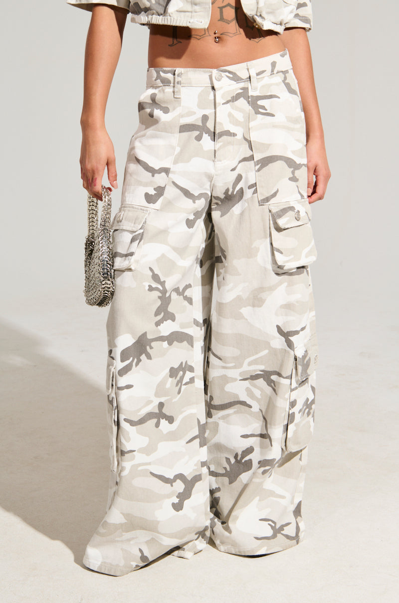 A person showing off the "YOUR UP NEXT CAMO CARGO PANT" in beige and white, designed with a high-rise fit and cargo pockets, accessorized with a small silver chain strap bag against a plain, light-colored background.