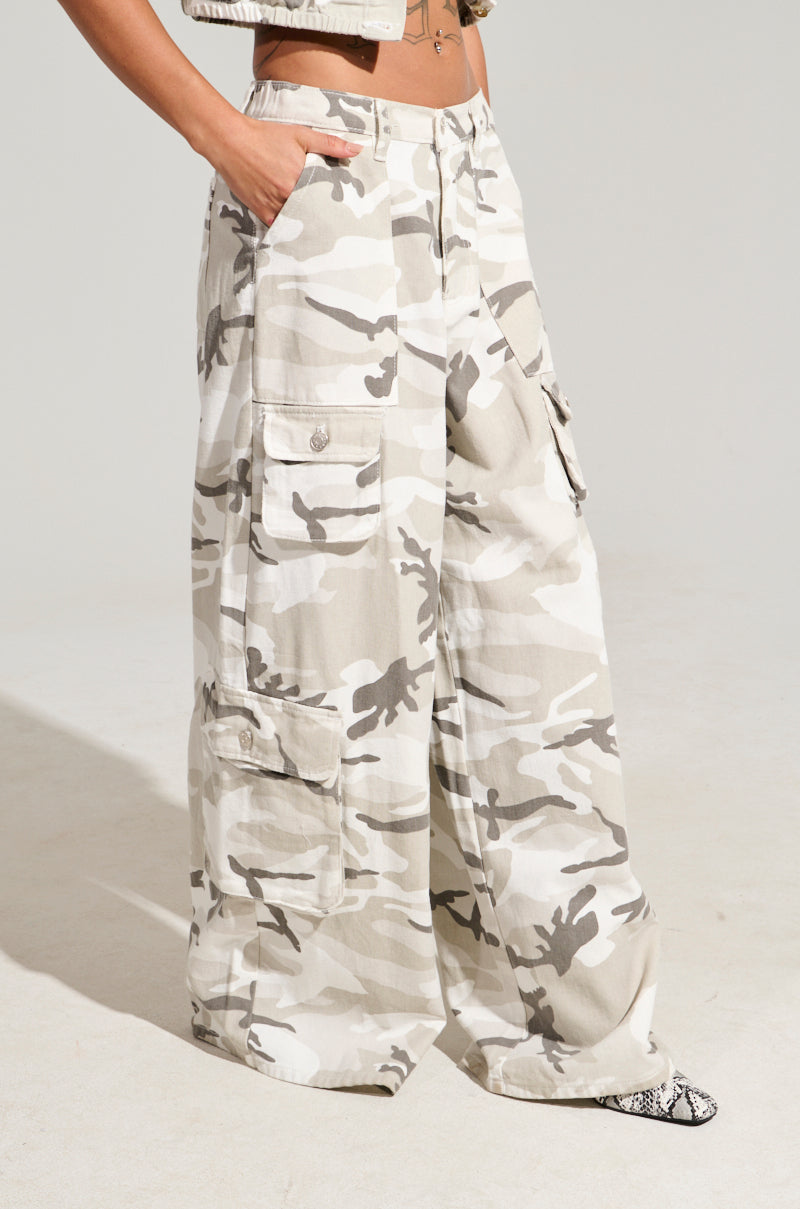 A person wearing the YOUR UP NEXT CAMO CARGO PANT, featuring a loose-fitting design in beige and white with large cargo pockets, stands against a plain background. They have their hands in the pockets of the pants and are also sporting a cropped jacket and snakeskin-patterned shoes.