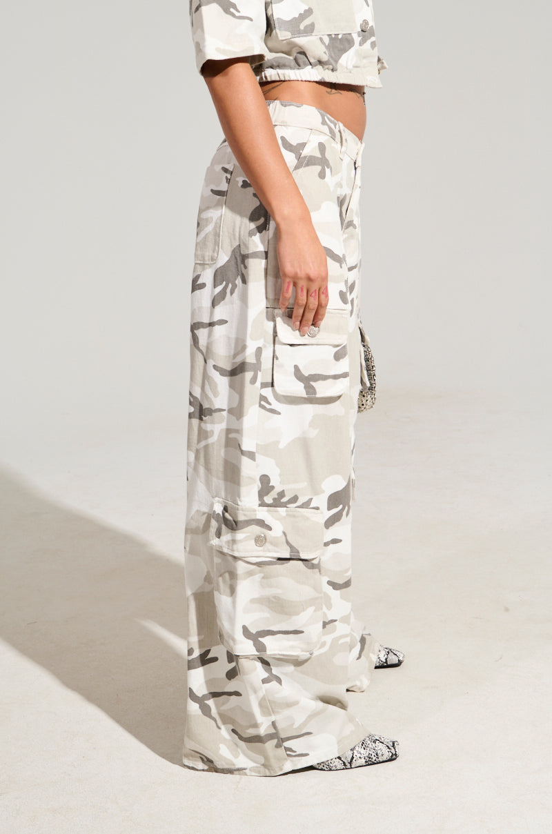 A person dressed in a beige and white camouflage-patterned outfit is showcased. The ensemble includes a short-sleeve top paired with the YOUR UP NEXT CAMO CARGO PANT, which features high-rise camo pants with cargo pockets. Their hand rests relaxed by their side as they stand against a plain background.