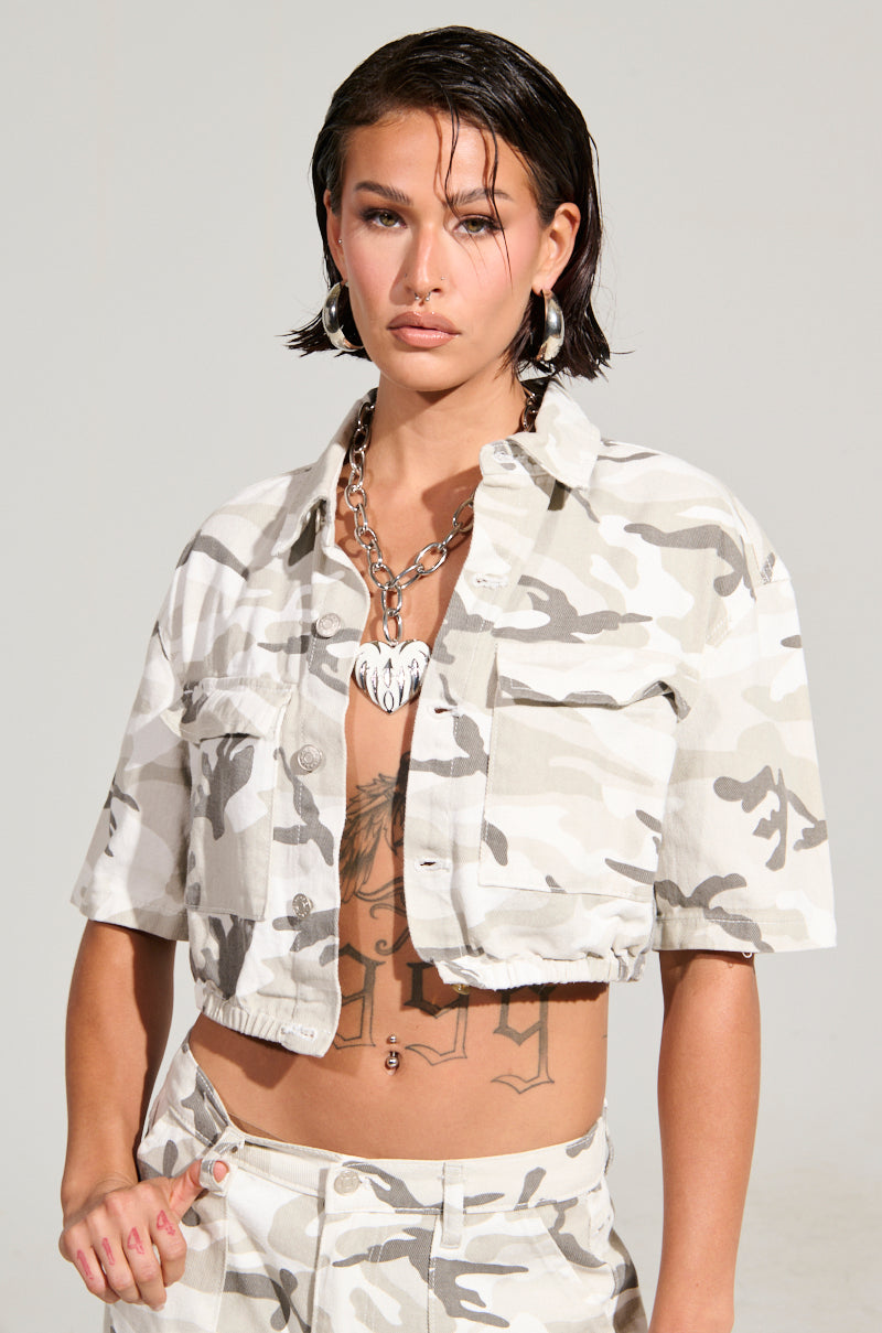 A person with short dark hair wears the YOURE UP NEXT CAMO BUTTON DOWN TOP, showcasing a cropped silhouette and matching elastic waistband pants. They have a tattoo on their stomach and a silver heart-shaped necklace. Their expression is serious as they stand against a plain background.