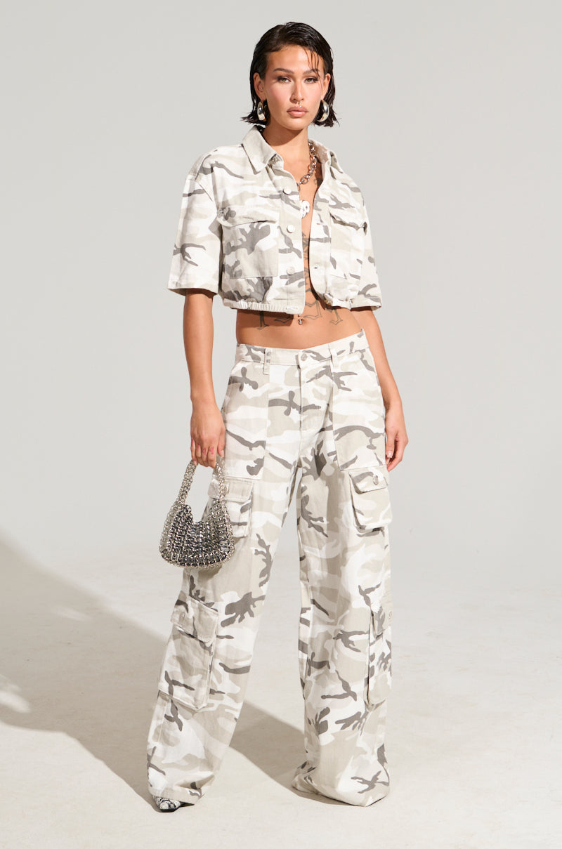 Dressed confidently in the "You're Up Next Camo Button Down Top," a person pairs it with pants featuring an elastic waistband. They complement their look with a small, beaded handbag and a striking necklace, creating a chic vibe against the plain, well-lit background.