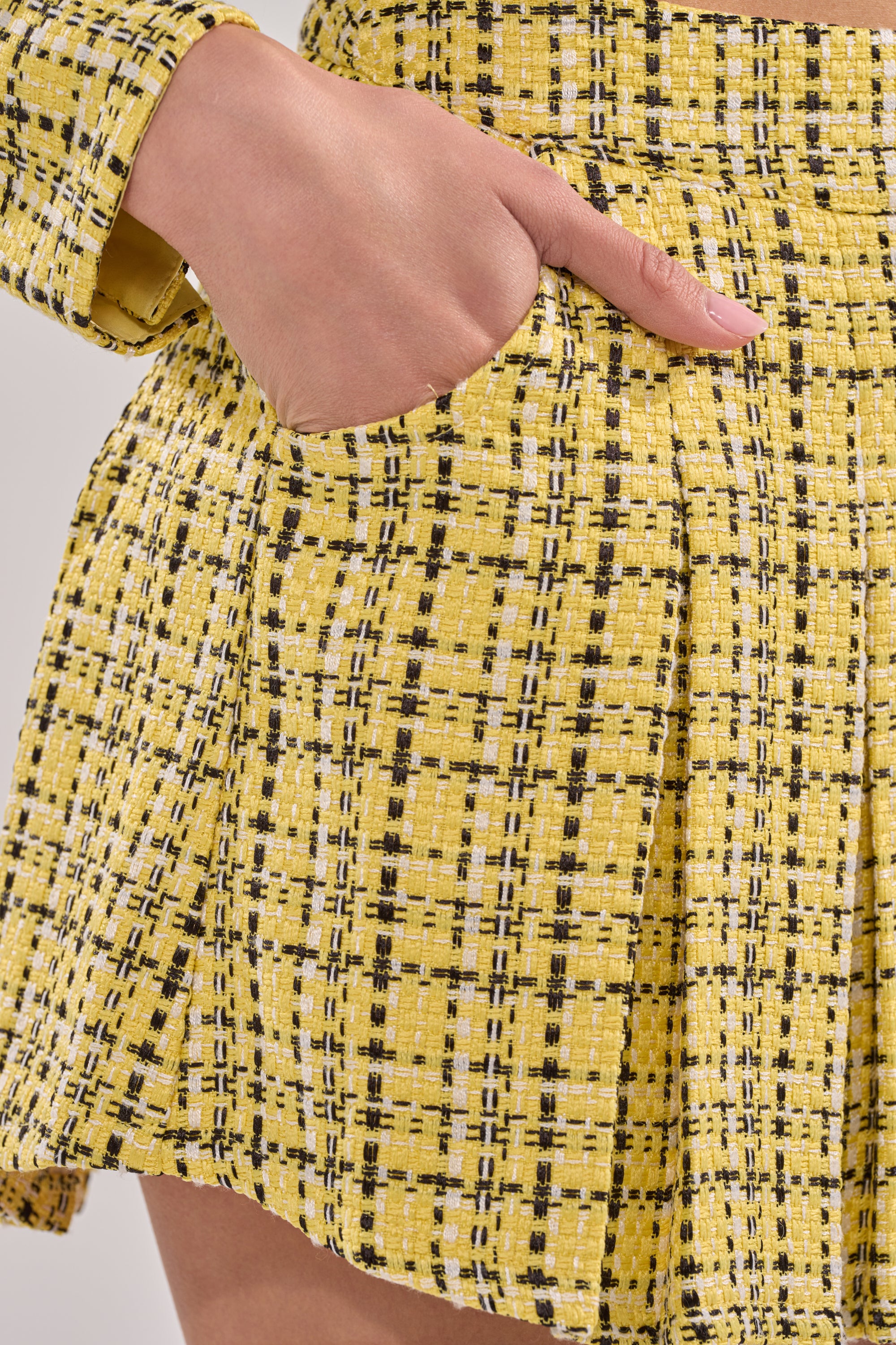 A close-up of a hand in the pocket of the FINN TWEED MINI SKIRT, featuring yellow woven checkered fabric and part of a matching jacket sleeve. 9