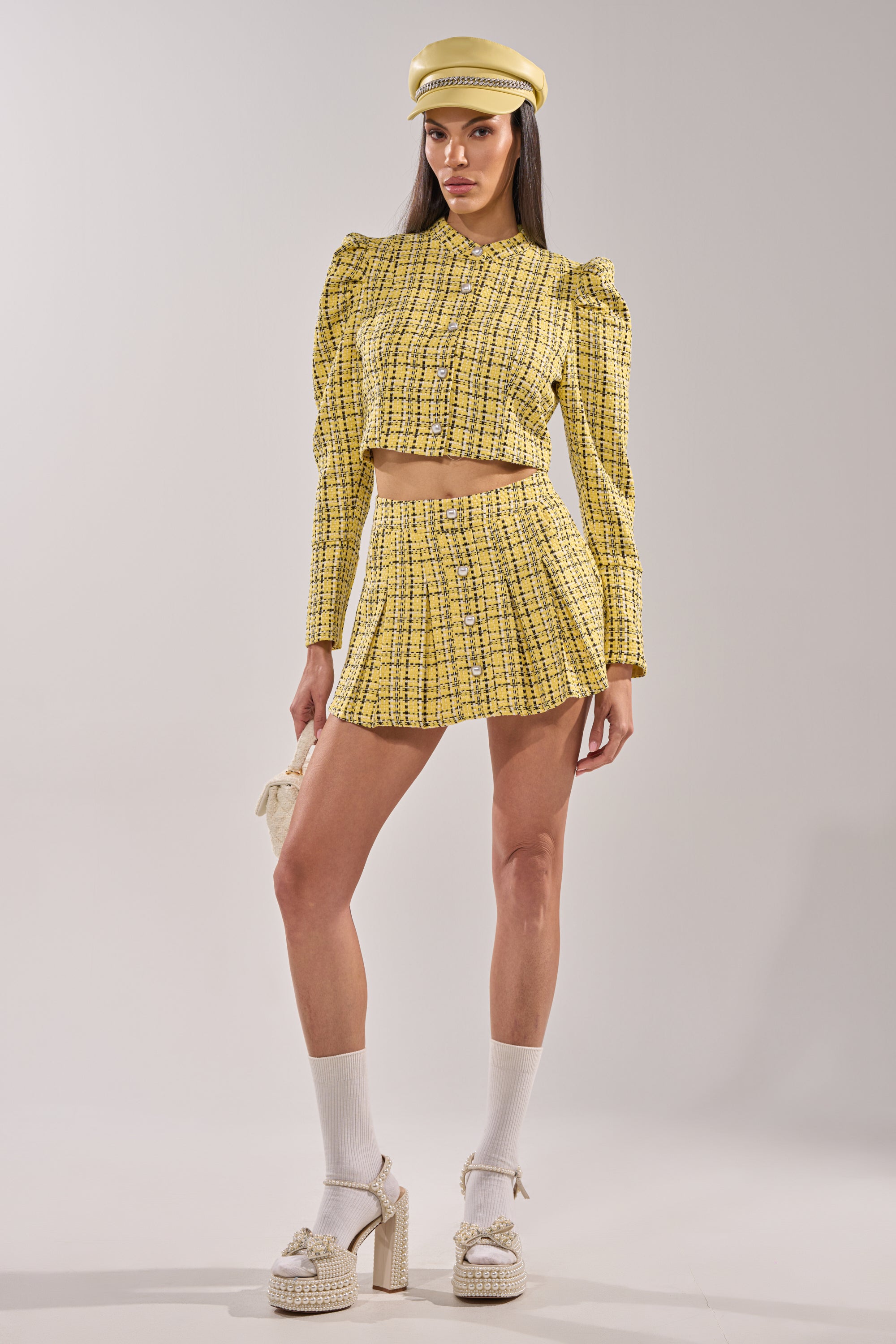 A woman wears the FINN TWEED MINI SKIRT—a yellow tweed mini skirt with buttons—paired with a matching plaid hat, a long-sleeve cropped jacket, white socks, chunky platform shoes, and holds a small purse against a plain background. 8