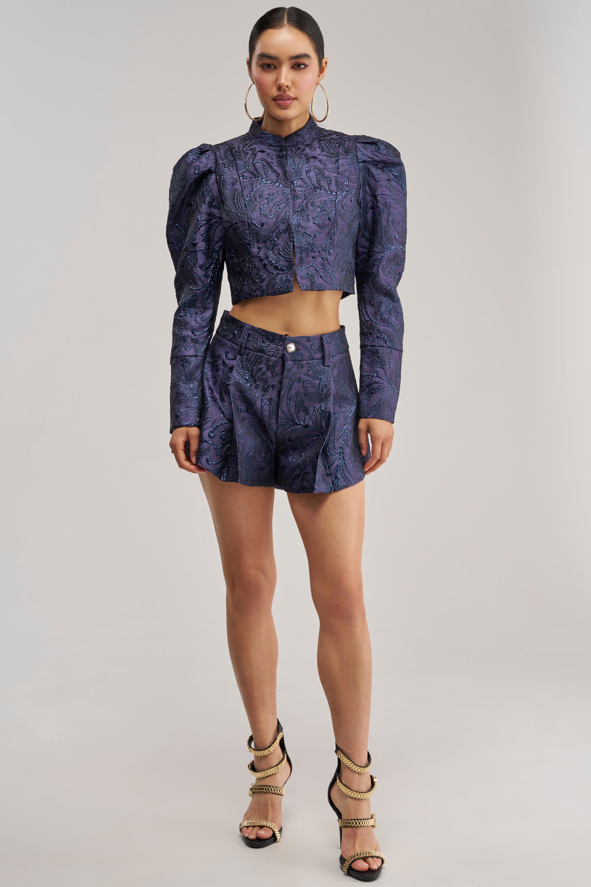 A woman wears the PENNY BROCADE SHORT IN NAVY, styled with a dark purple textured crop top with puffed shoulders, strappy high heels, and hoop earrings. She stands facing forward against a plain light background.