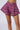 A person is wearing the high-waisted EFFIE TWEED SHORTS, crafted from a vibrant, multicolored tweed fabric. The chic tweed material incorporates a pleated design and slightly flares at the sides, giving it a skirt-like appearance. The background is plain and light-colored.