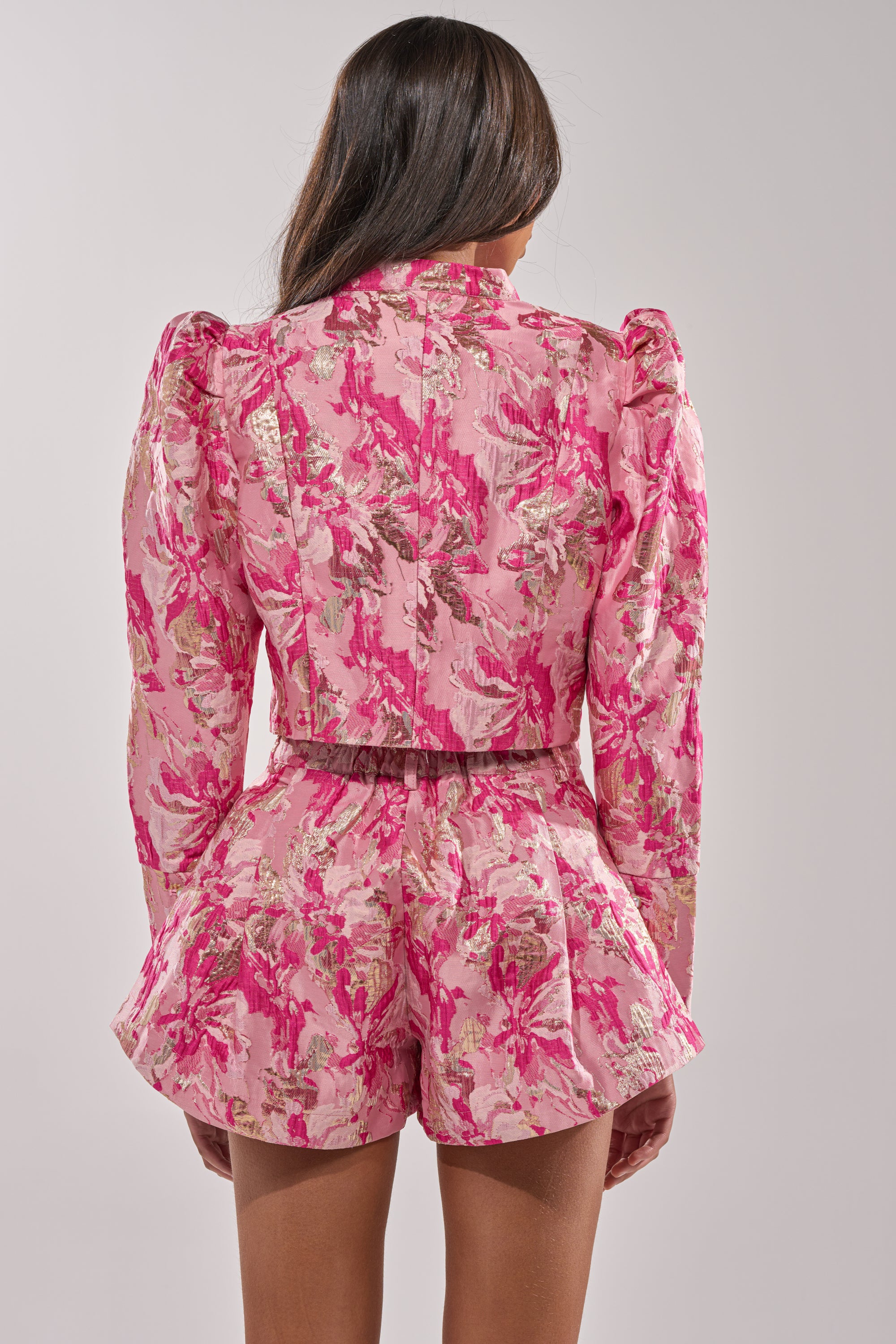 A woman with long brown hair, seen from the back, wears the POPPY BROCADE BLAZER and matching high-waisted shorts in pink floral jacquard with puffed shoulders, standing against a plain light gray background. 3