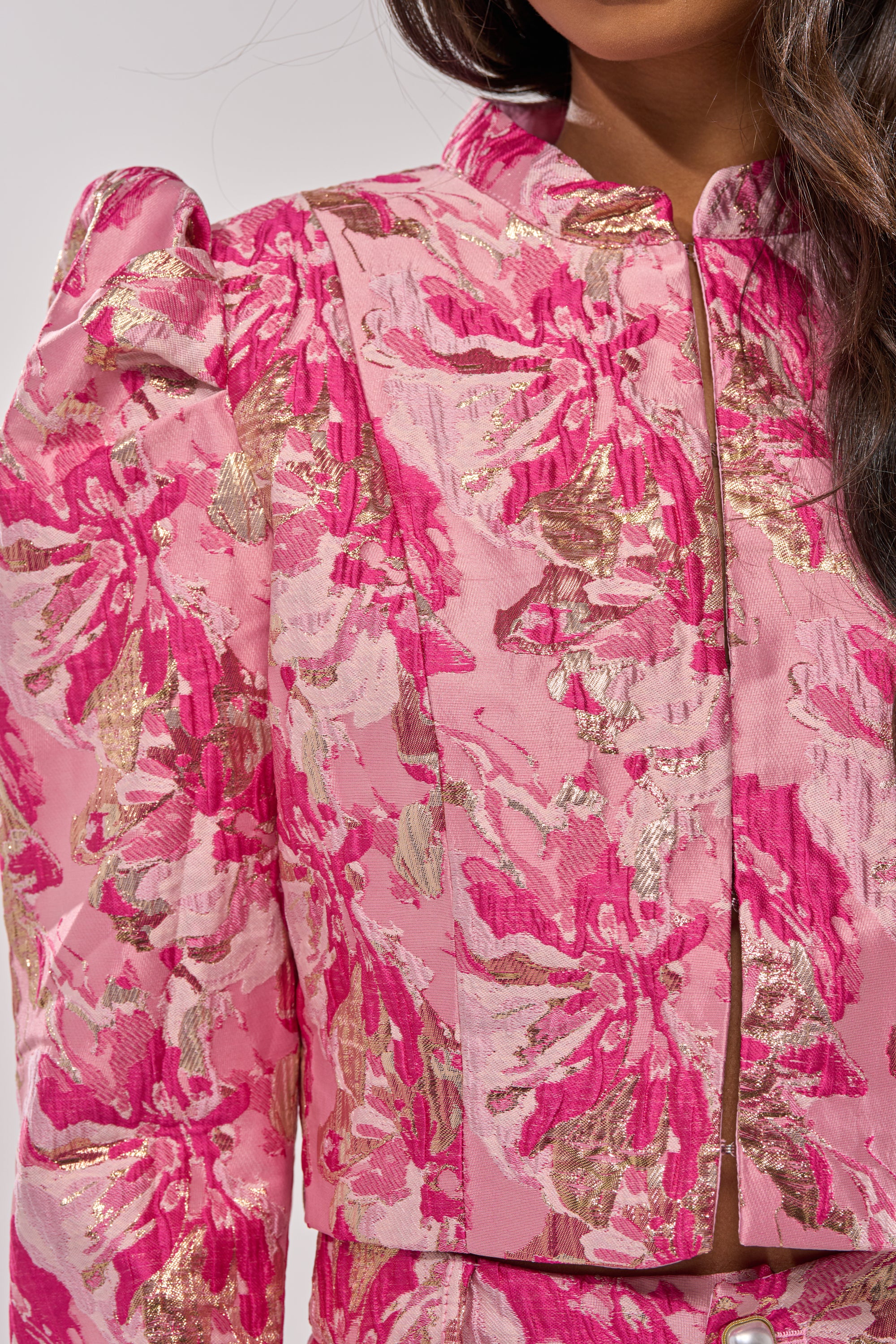 A woman models the POPPY BROCADE BLAZER, featuring pink and gold floral jacquard, puffed shoulders, a round neckline, and a vibrant metallic sheen. The photo is cropped to display only her upper body. 5