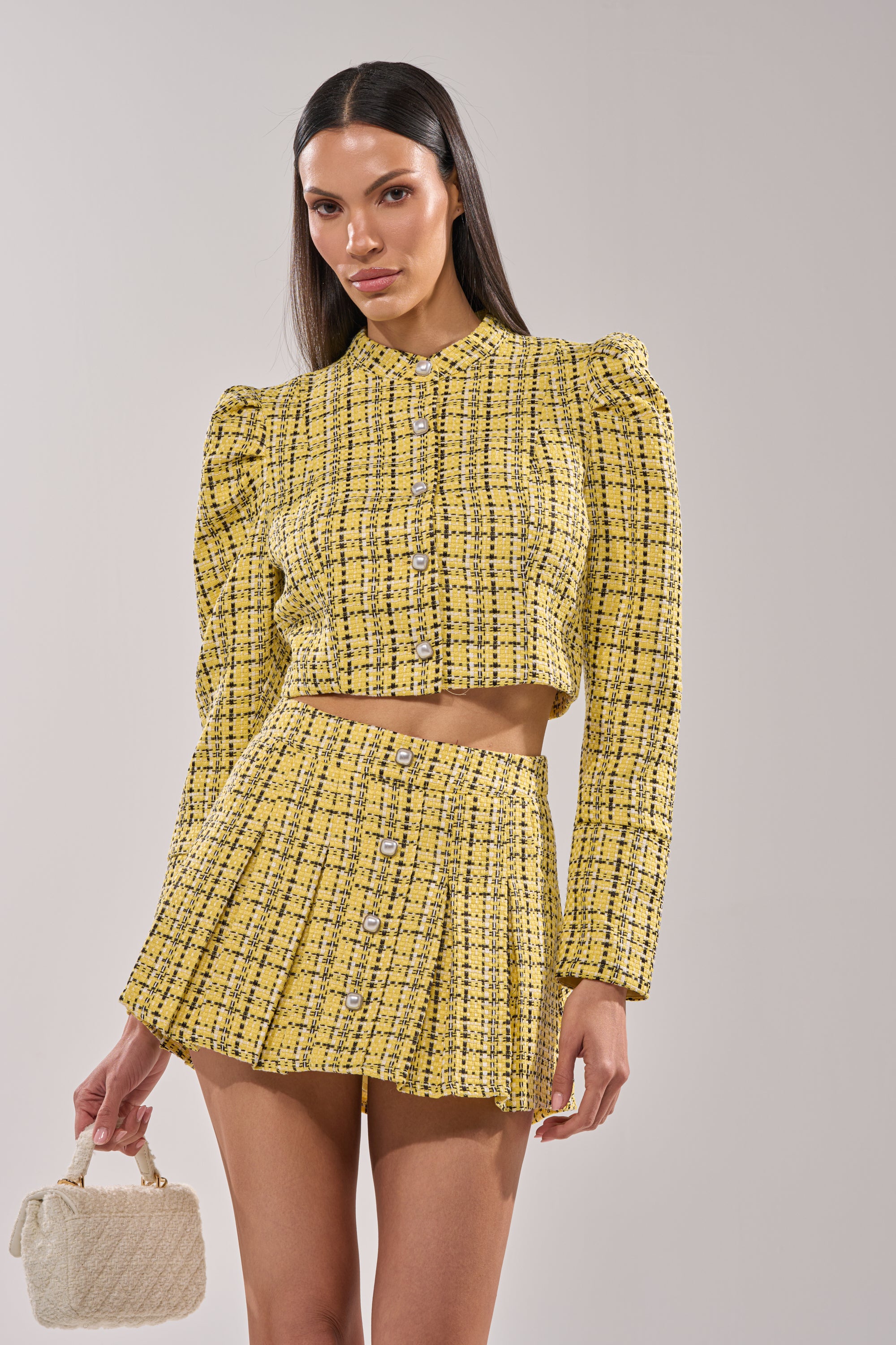 A woman poses against a plain light background wearing the FINN TWEED BLAZER in yellow, featuring pearl buttons, paired with a matching high-waisted pleated mini skirt and holding a small white textured handbag. 0