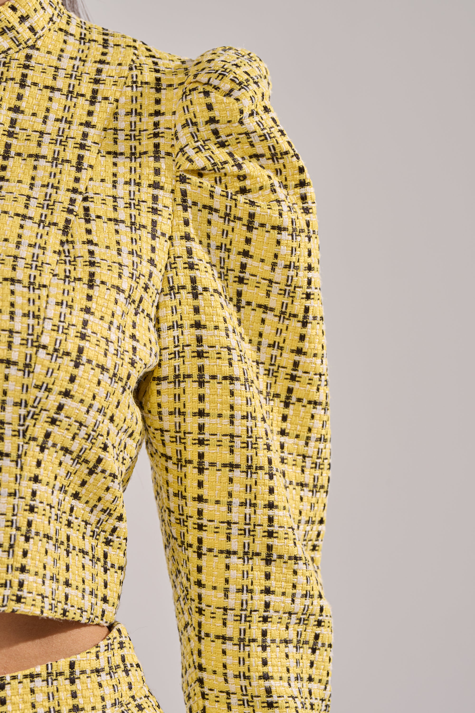 Close-up of a person wearing the FINN TWEED BLAZER, featuring yellow, black, and white plaid, puffed shoulders, pearl buttons, and a cropped cut against a neutral gray background. 9