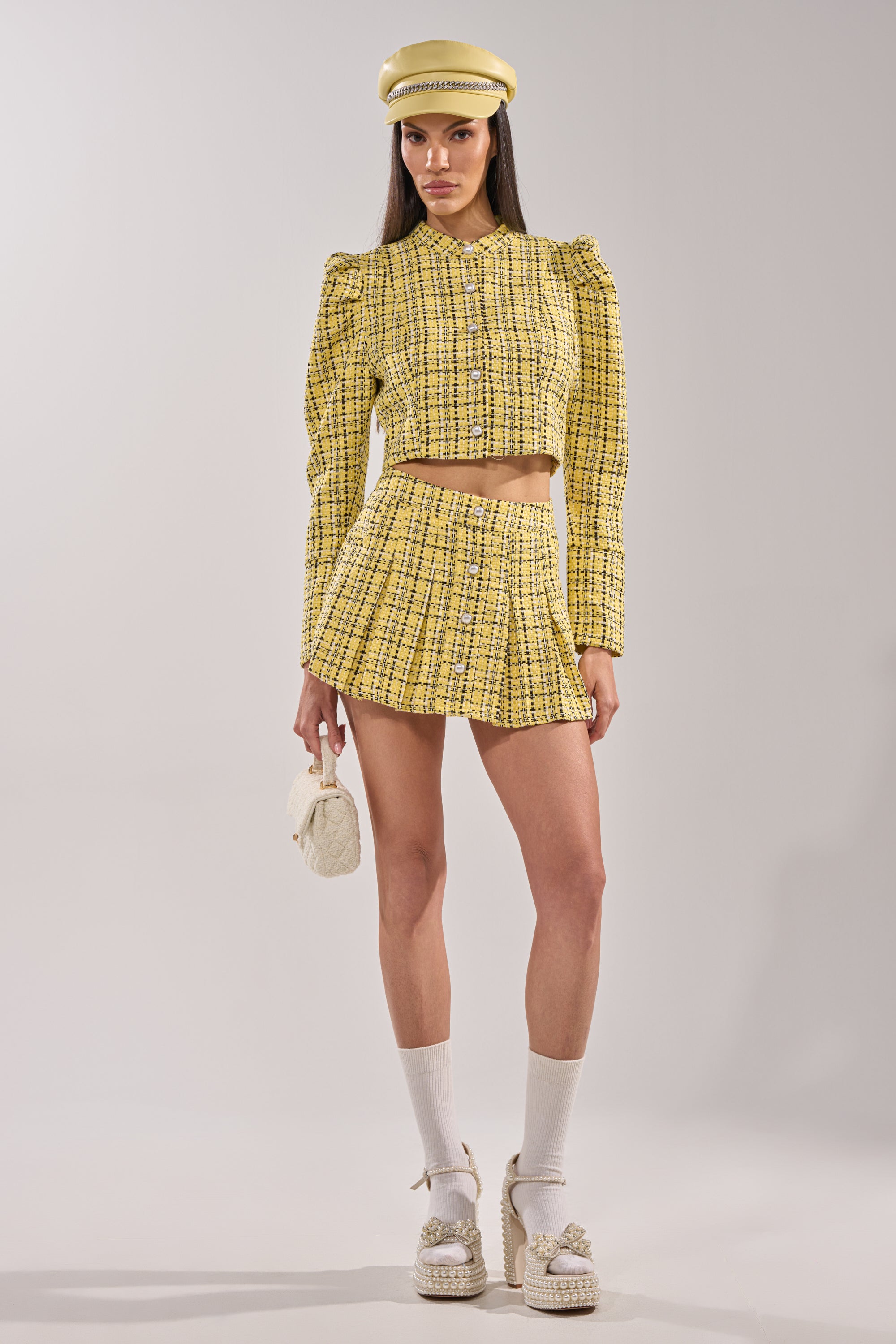 A woman in a yellow plaid matching set with the FINN TWEED BLAZER featuring pearl buttons, a mini skirt, white socks, platform sandals, a yellow cap, and a small white bag stands against a plain light background. 10