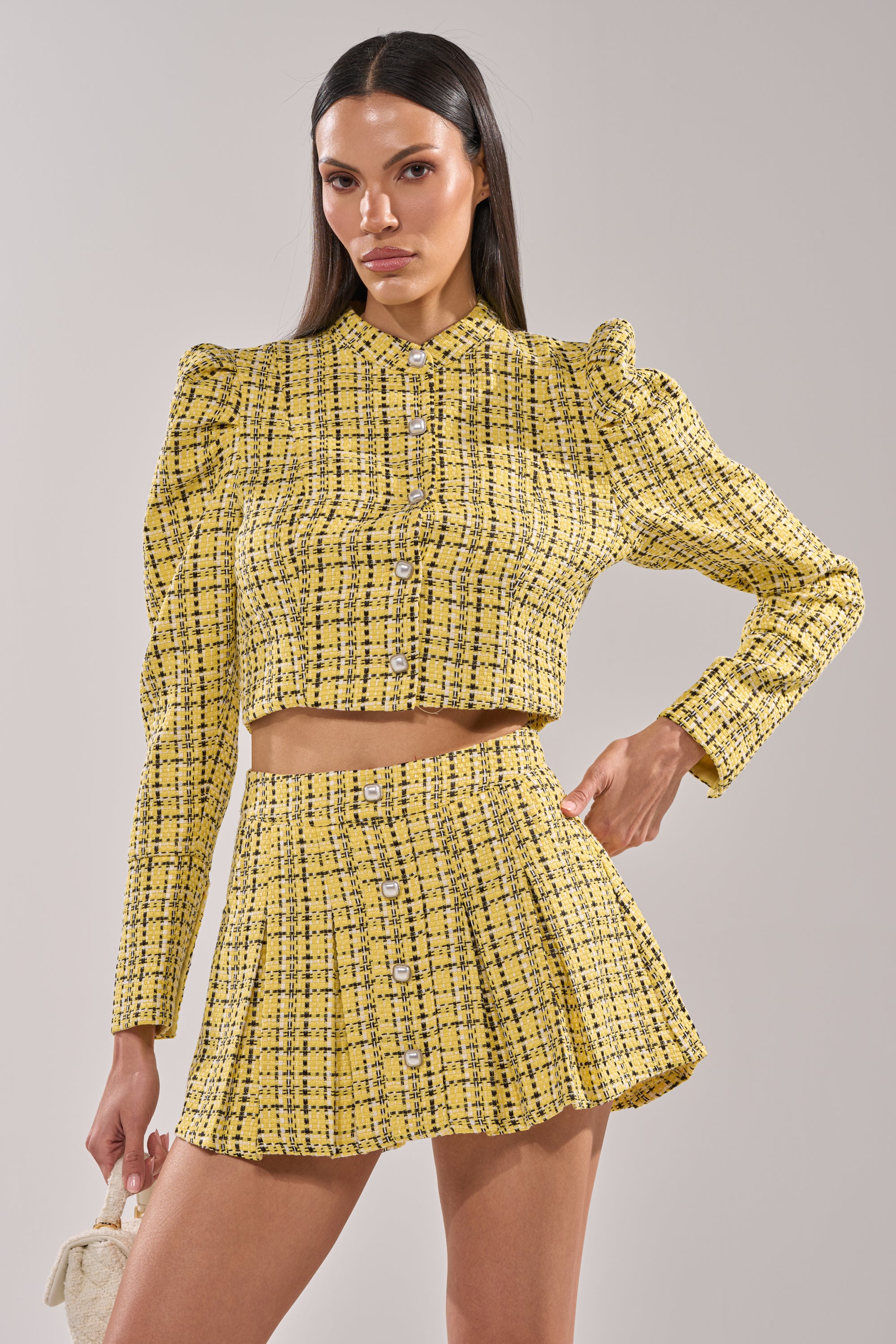 A woman poses confidently in the FINN TWEED BLAZER, a yellow plaid, button-up, long-sleeve crop top with pearl buttons, paired with a high-waisted pleated mini skirt. She sports long dark hair and carries a small white purse. 1