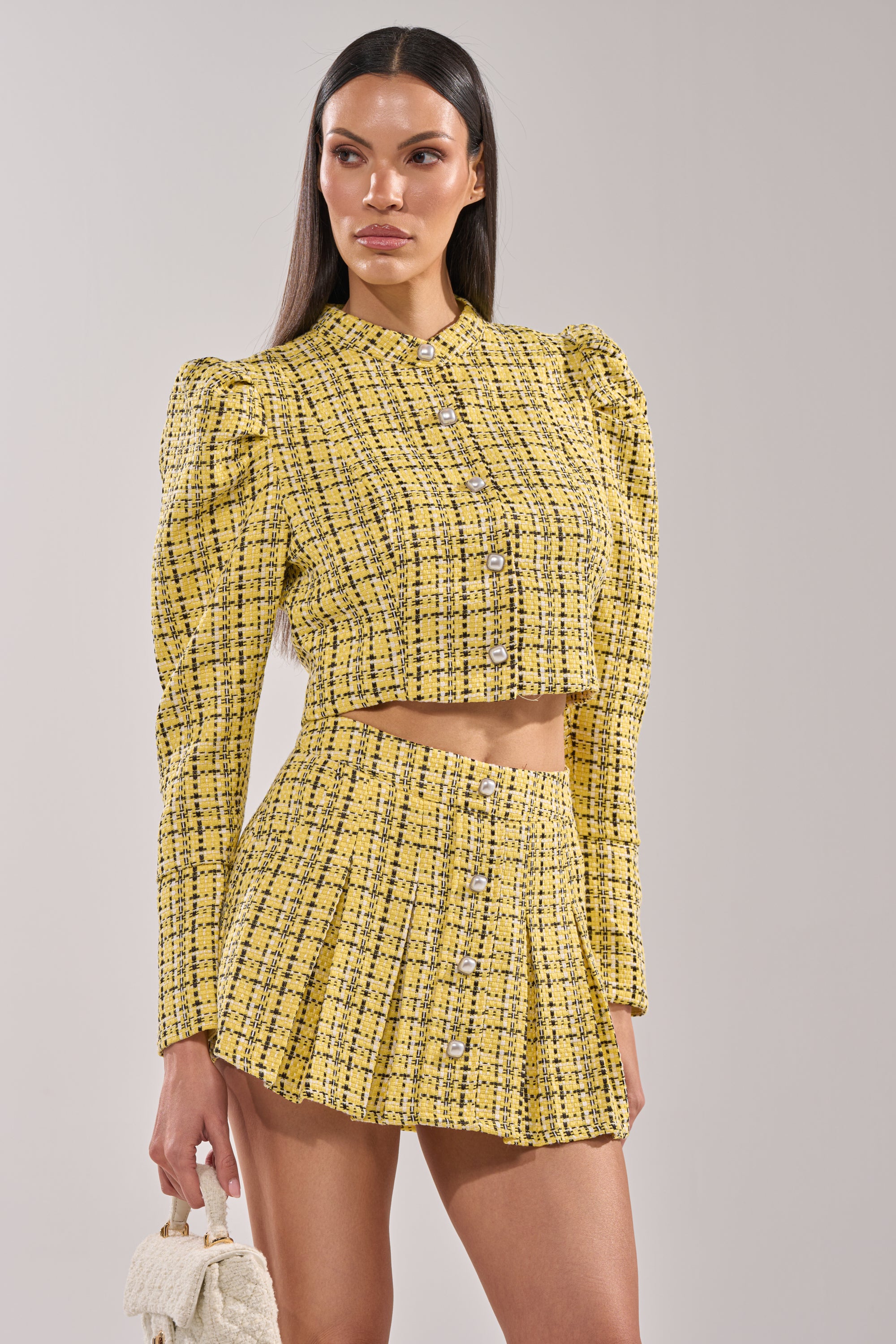 A woman wears the FINN TWEED BLAZER, a yellow plaid, long-sleeve crop top with pearl buttons, paired with a matching high-waisted mini skirt. She carries a small textured white handbag and stands against a plain background. 2