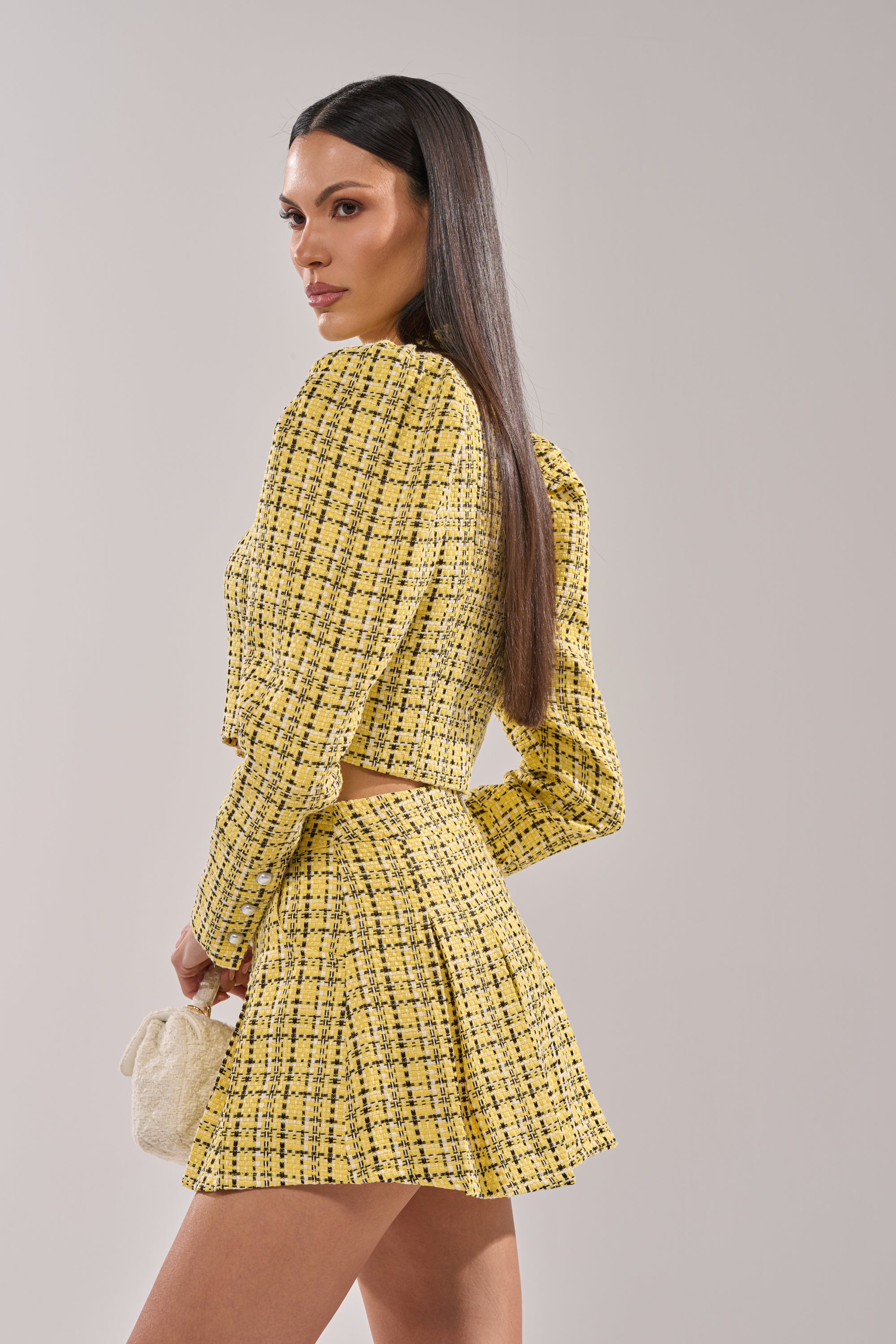 A woman poses against a light gray background wearing the FINN TWEED BLAZER in yellow plaid with pearl buttons, paired with a matching pleated mini skirt and holding a small cream textured handbag. 3