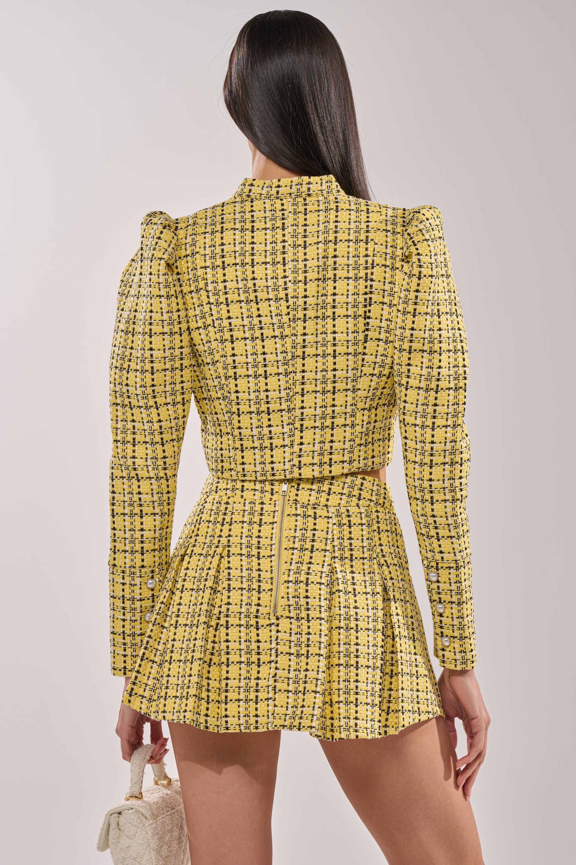 A person with long, straight dark hair is seen from the back wearing the FINN TWEED BLAZER—a yellow tweed cropped blazer with pearl buttons—paired with a matching high-waisted plaid skirt and holding a small textured beige handbag. 4