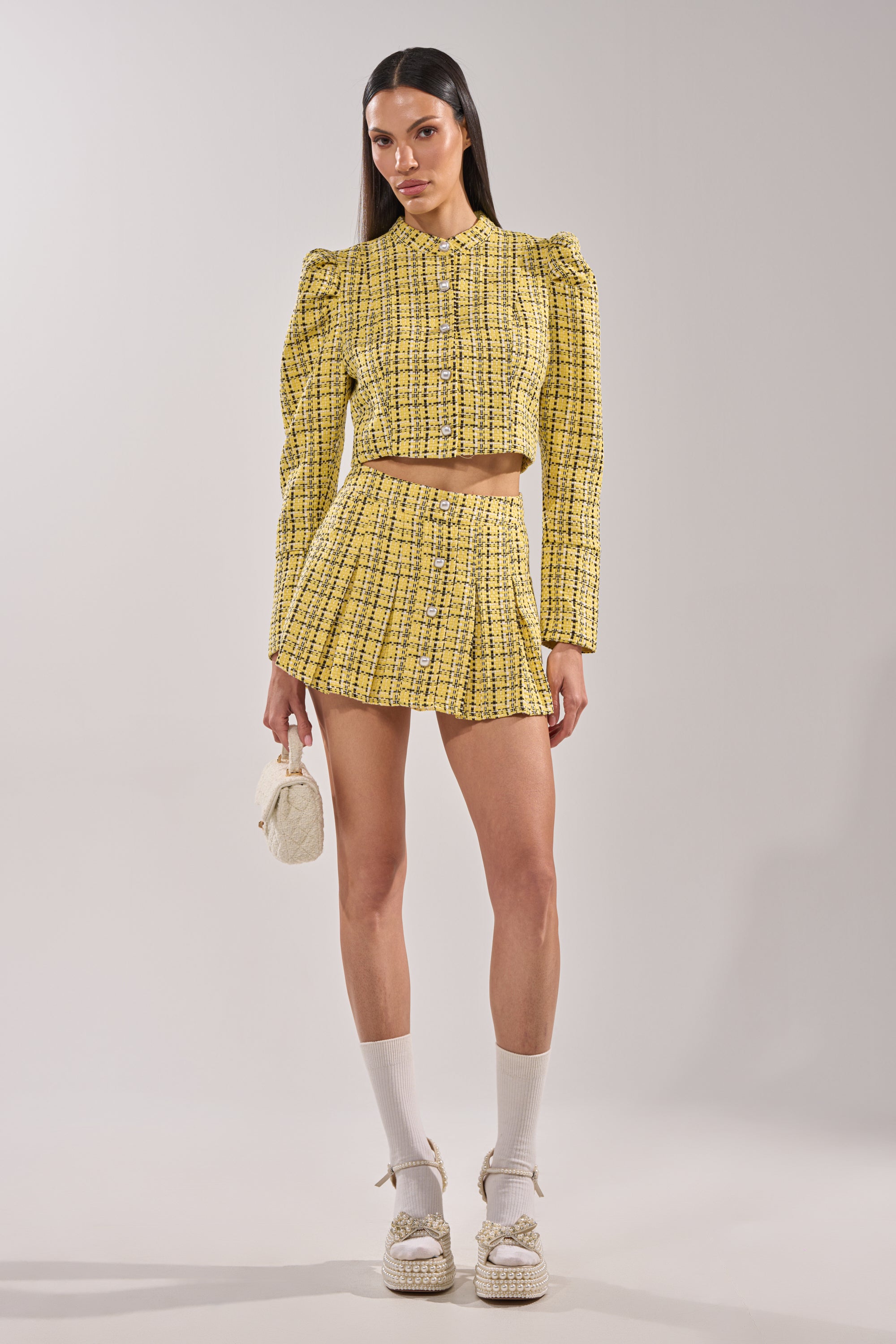 A woman stands in a studio wearing the FINN TWEED BLAZER in yellow plaid with pearl buttons and puffed shoulders, styled with a matching pleated mini skirt, a small white bag, white socks, and white platform sandals. 6