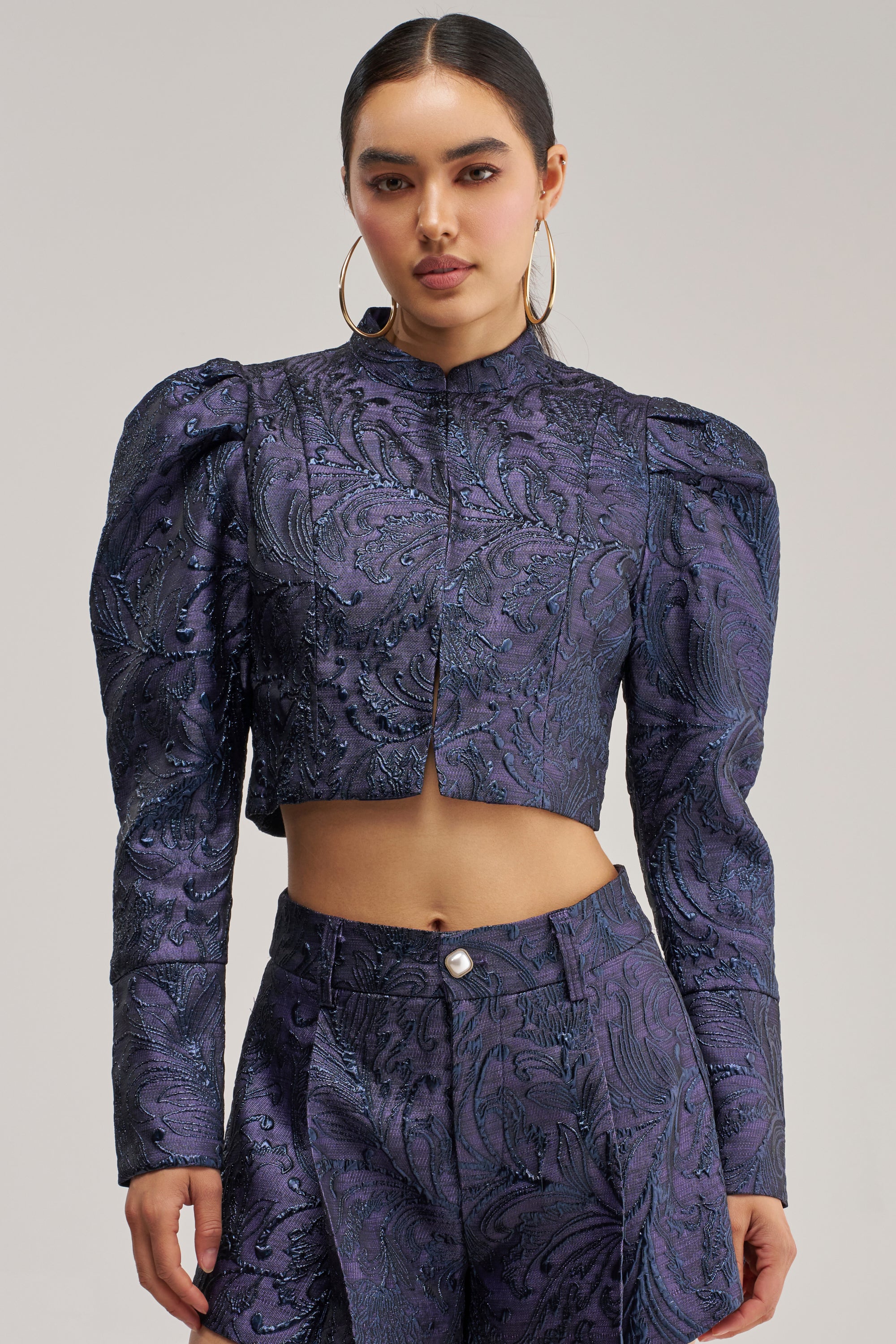 A woman wears the PENNY BROCADE BLAZER IN NAVY, a cropped blazer with structured shoulders that gives her look a modern edge. She pairs it with high-waisted pants featuring a paisley pattern, hoop earrings, and has her hair pulled back.
