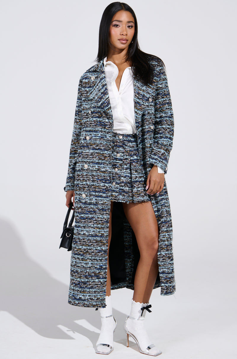 A woman with long black hair models a multicolored tweed coat featuring embossed silver buttons over the ALZURA TWEED MINI SKIRT and a white blouse. She completes her look with white high heels and holds a small black handbag, standing against a plain background.