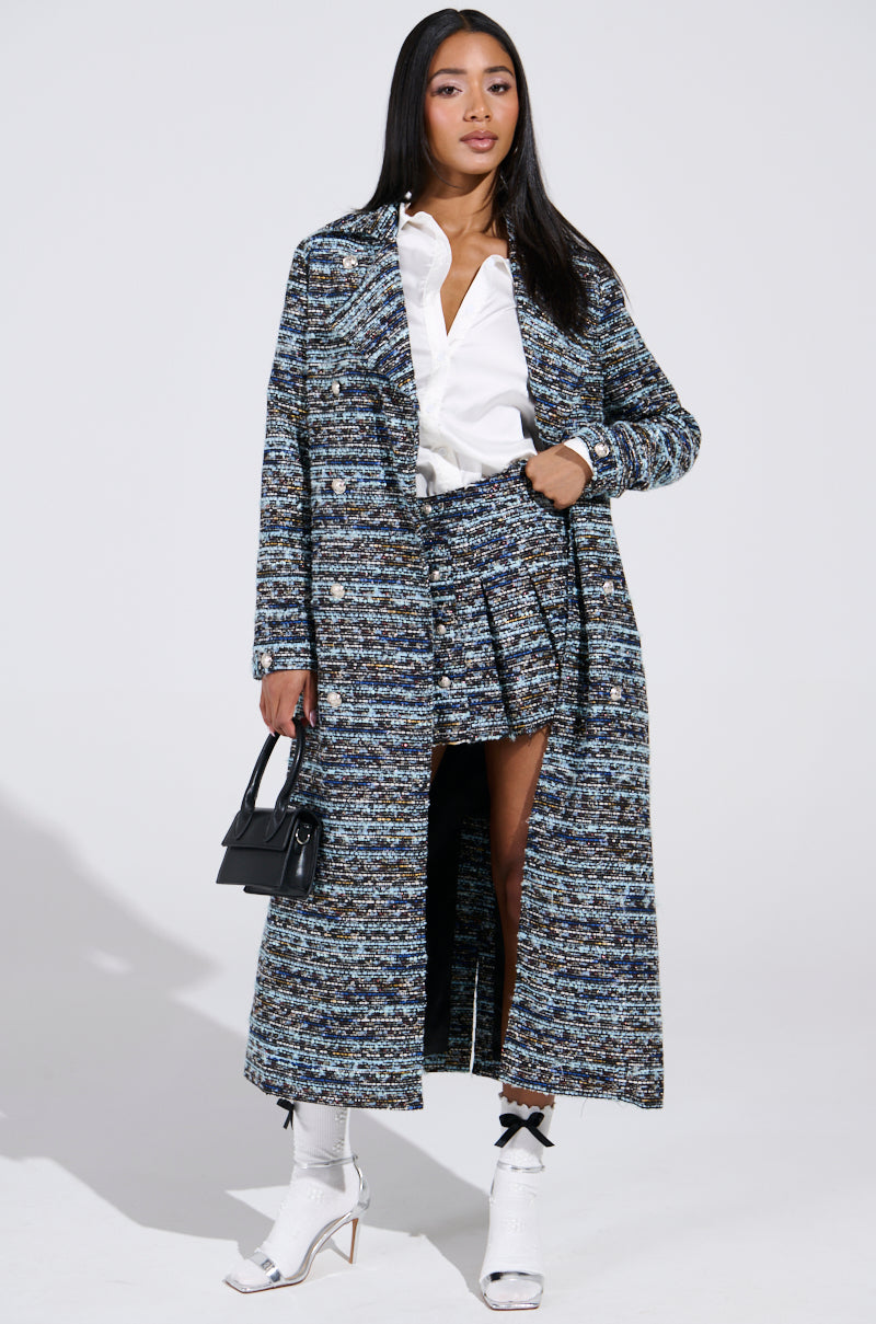 A woman poses confidently in a stylish blue and gray textured long coat with embossed silver buttons, a matching ALZURA TWEED MINI SKIRT, and a white blouse. She pairs it with white ankle boots, carries a small black handbag, all against a plain white background.