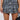 A person in an ALZURA Tweed Mini Skirt, featuring a blue and black check with embossed silver buttons, along with a white blouse and a small black handbag, stands against a light background.