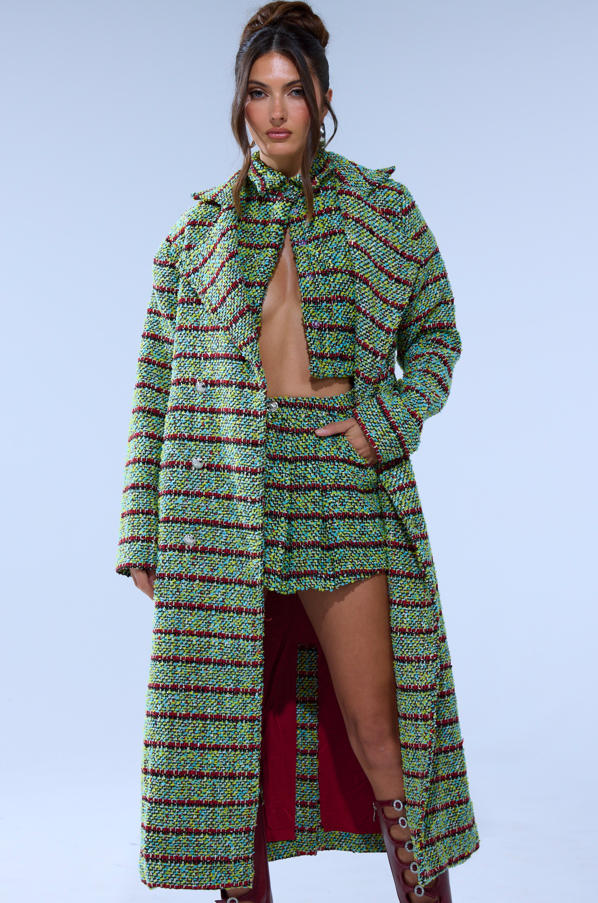A woman poses confidently in the IT'S GIVING TWEED MINI SKIRT, styled with a matching green, black, and pink plaid tweed crop top and long coat. Her burgundy lace-up boots pop against the plain light blue background.