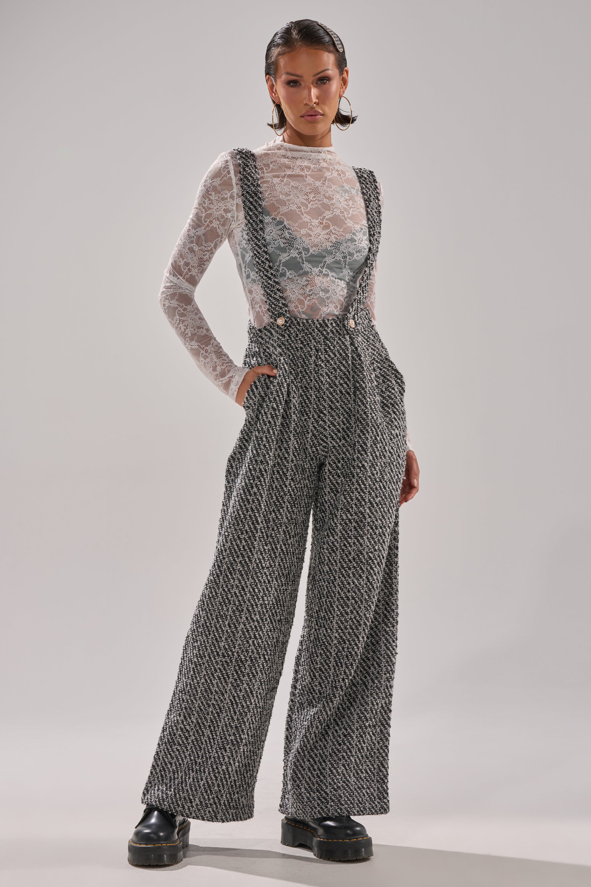 A woman stands confidently in ALESSANDRA TWEED OVERALLS with pearl button straps, paired with a sheer white lace long-sleeve top and black platform shoes against a plain light background.