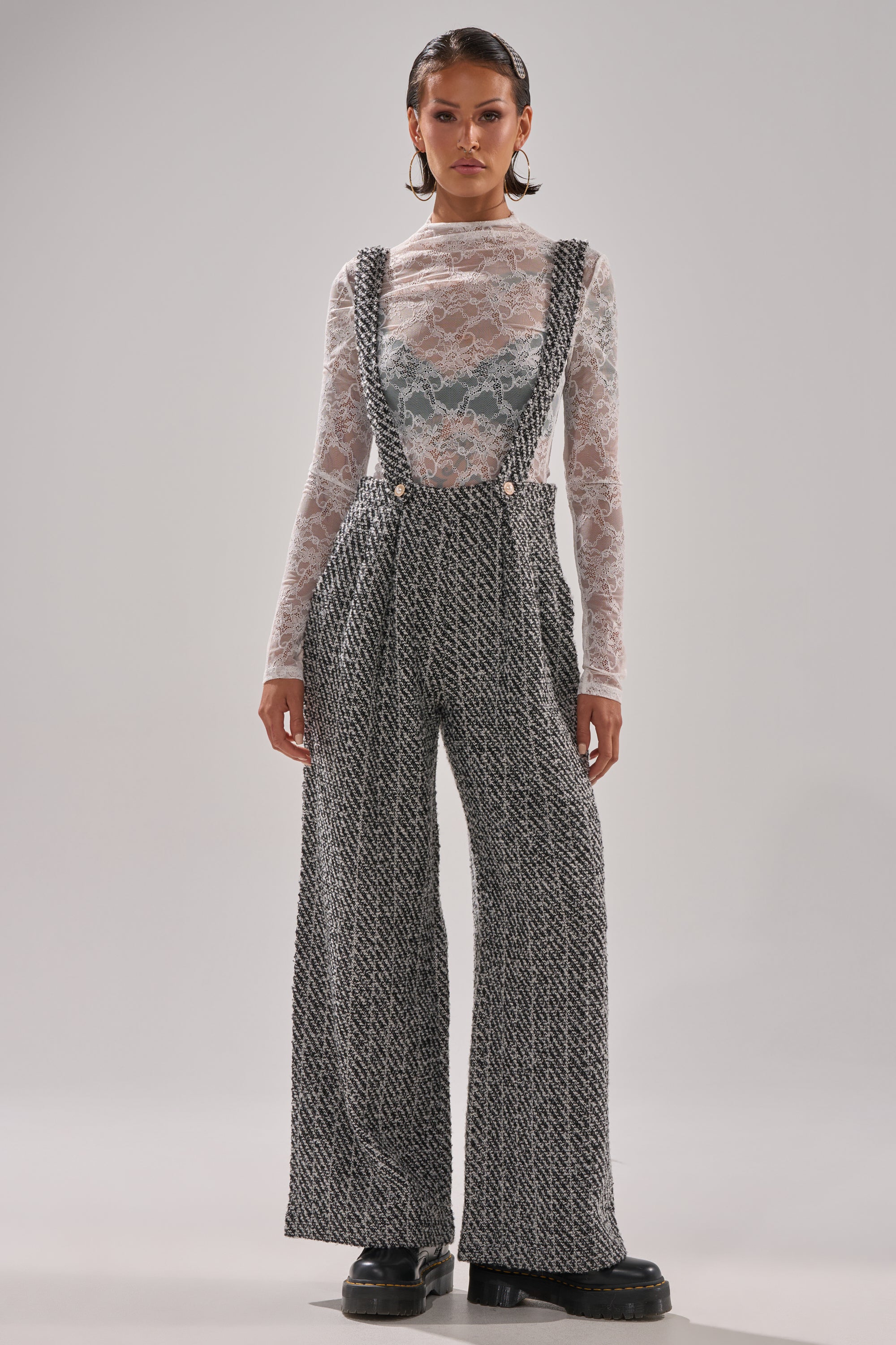 A woman faces forward wearing the ALESSANDRA TWEED OVERALLS, featuring pearl button straps over a sheer white lace long-sleeve top, paired with black chunky platform shoes.
