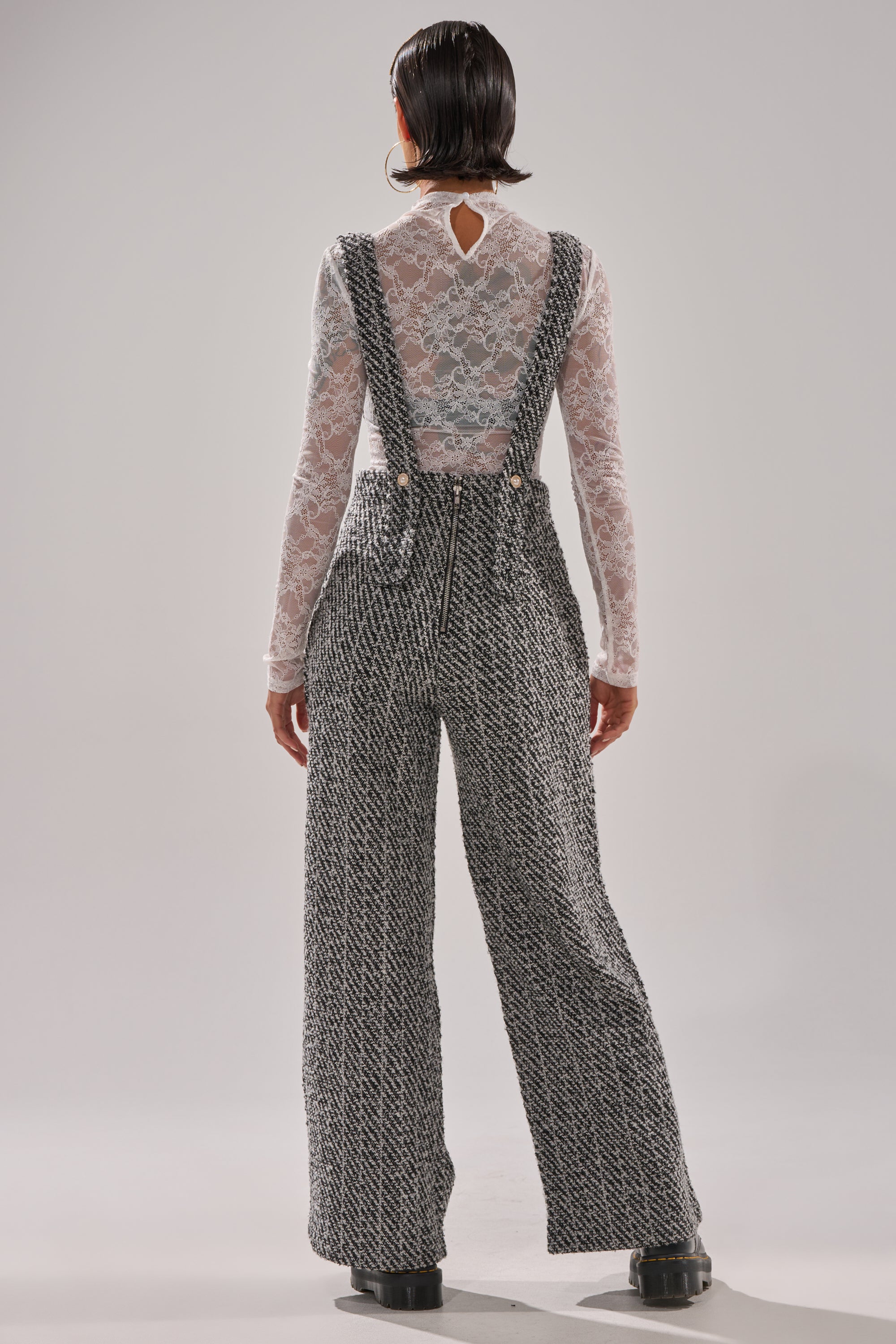 A person faces away, wearing ALESSANDRA TWEED OVERALLS—wide-leg gray and white textured overalls with suspenders—over a sheer lace top, styled with short, sleek hair and black platform shoes.
