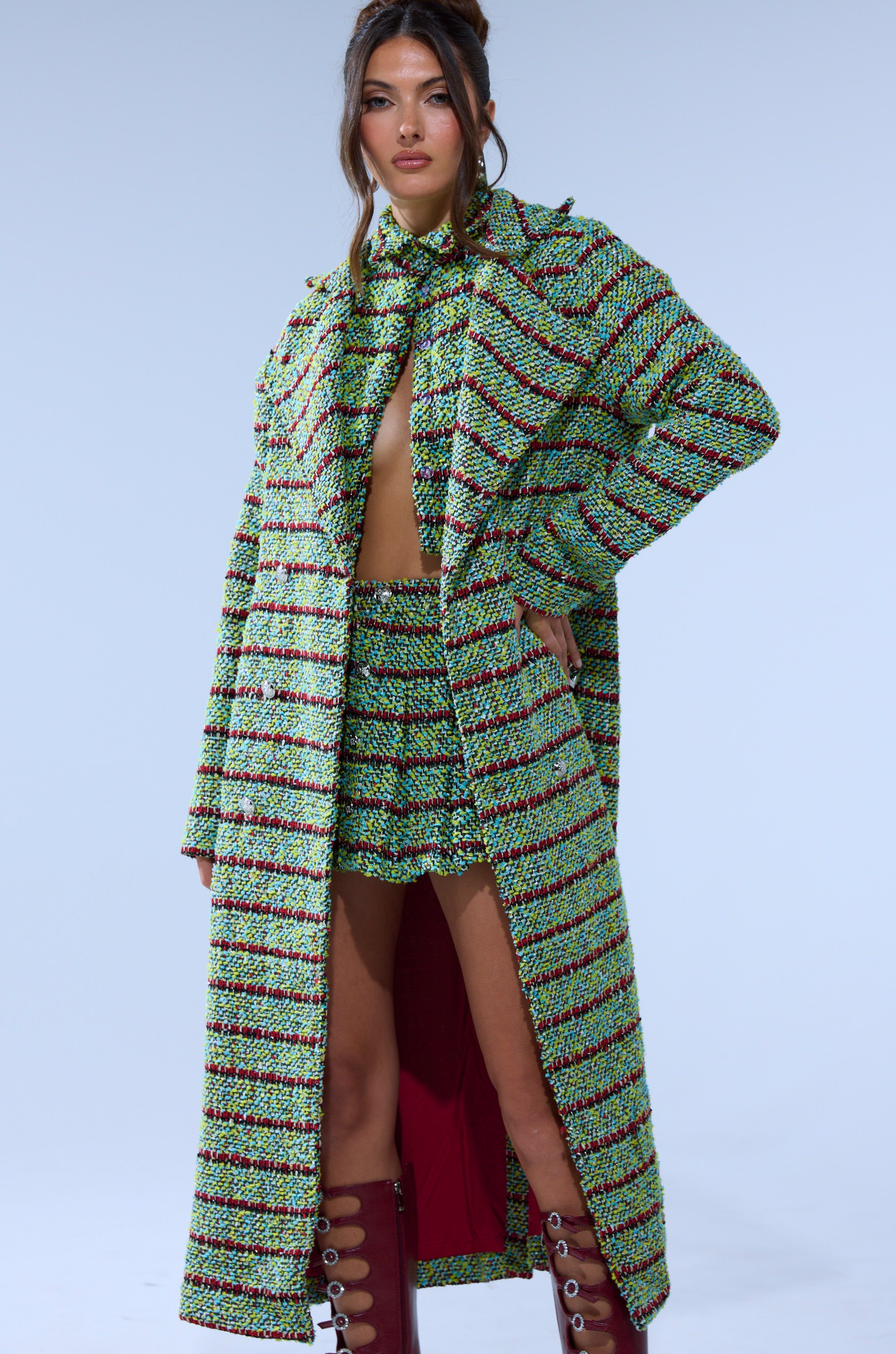 A woman poses in the IT'S GIVING LUCK TWEED TRENCH over a matching skirt and crop top, styled with burgundy lace-up knee-high boots against a light blue background.