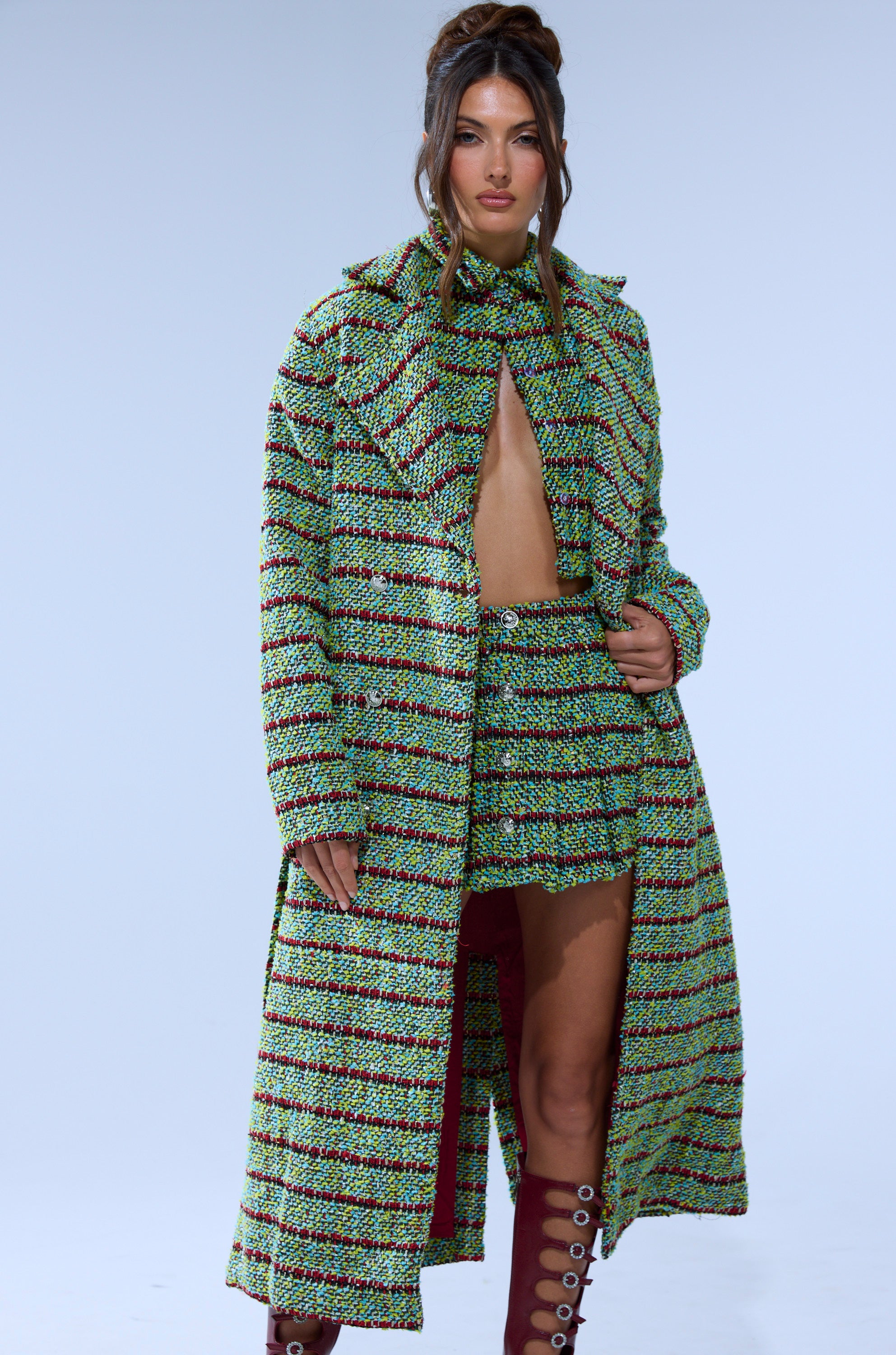 A woman models the IT'S GIVING LUCK TWEED TRENCH over a matching mini skirt and bandeau top, paired with knee-high burgundy boots, standing against a plain light background.