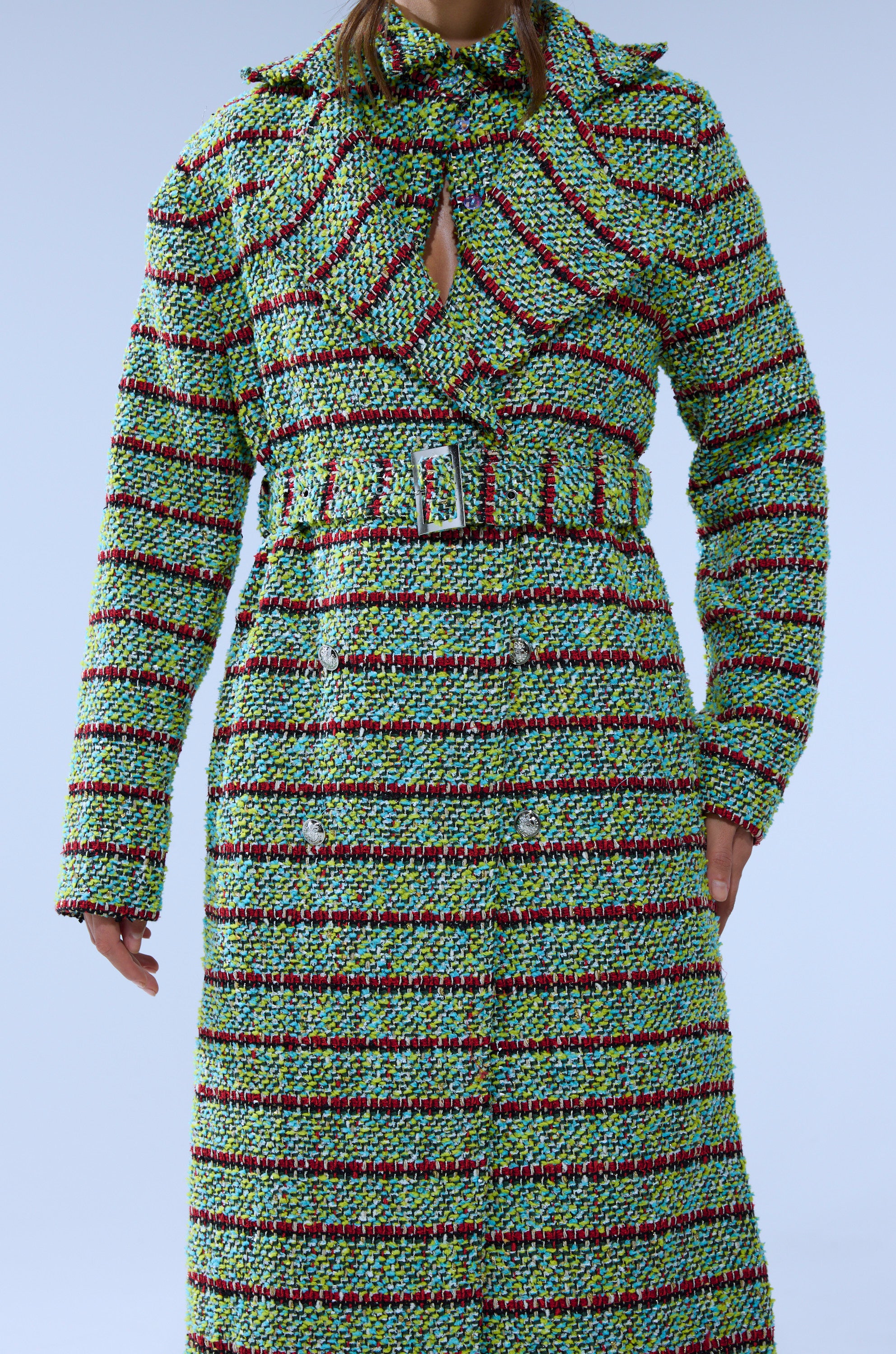 A person wears the IT'S GIVING LUCK TWEED TRENCH, a textured, belted coat with green, red, and black horizontal stripes, standing against a pale blue background with their head partially out of frame.