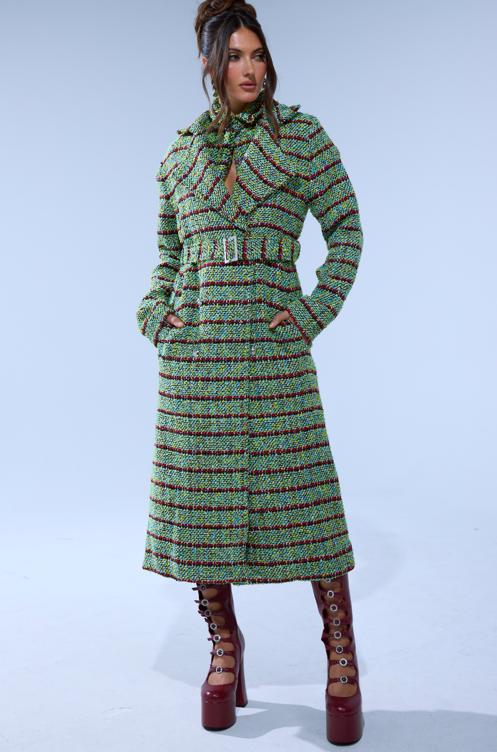 A woman wears the IT'S GIVING LUCK TWEED TRENCH, a longline multicolor tweed coat with green, red, and black stripes. She styles it with maroon platform boots and a matching scarf, posing against a light blue background.