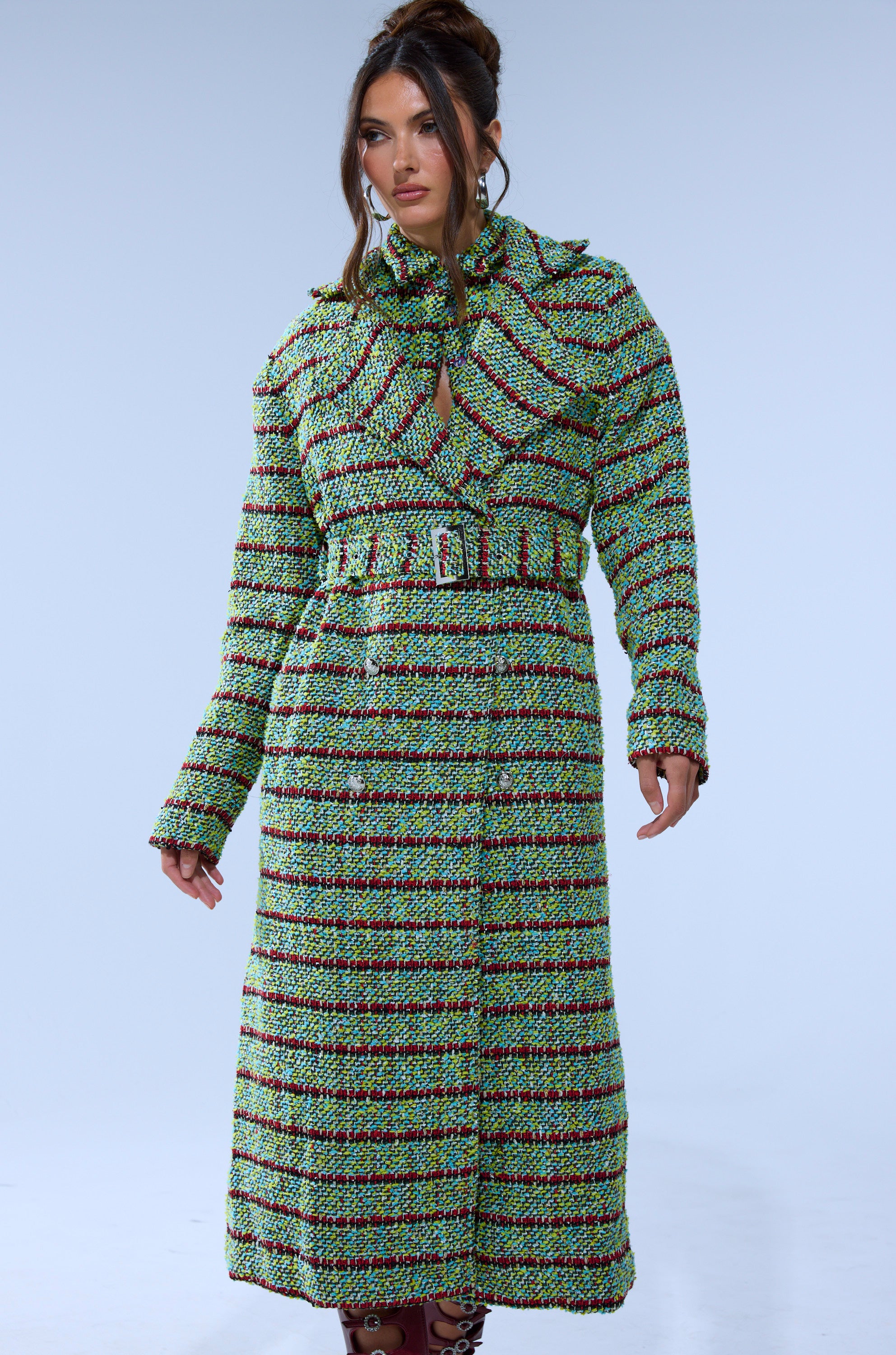 A woman stands against a light blue background wearing the IT'S GIVING LUCK TWEED TRENCH, featuring a longline silhouette, green and red stripes, belted waist, high collar, and keyhole neckline.