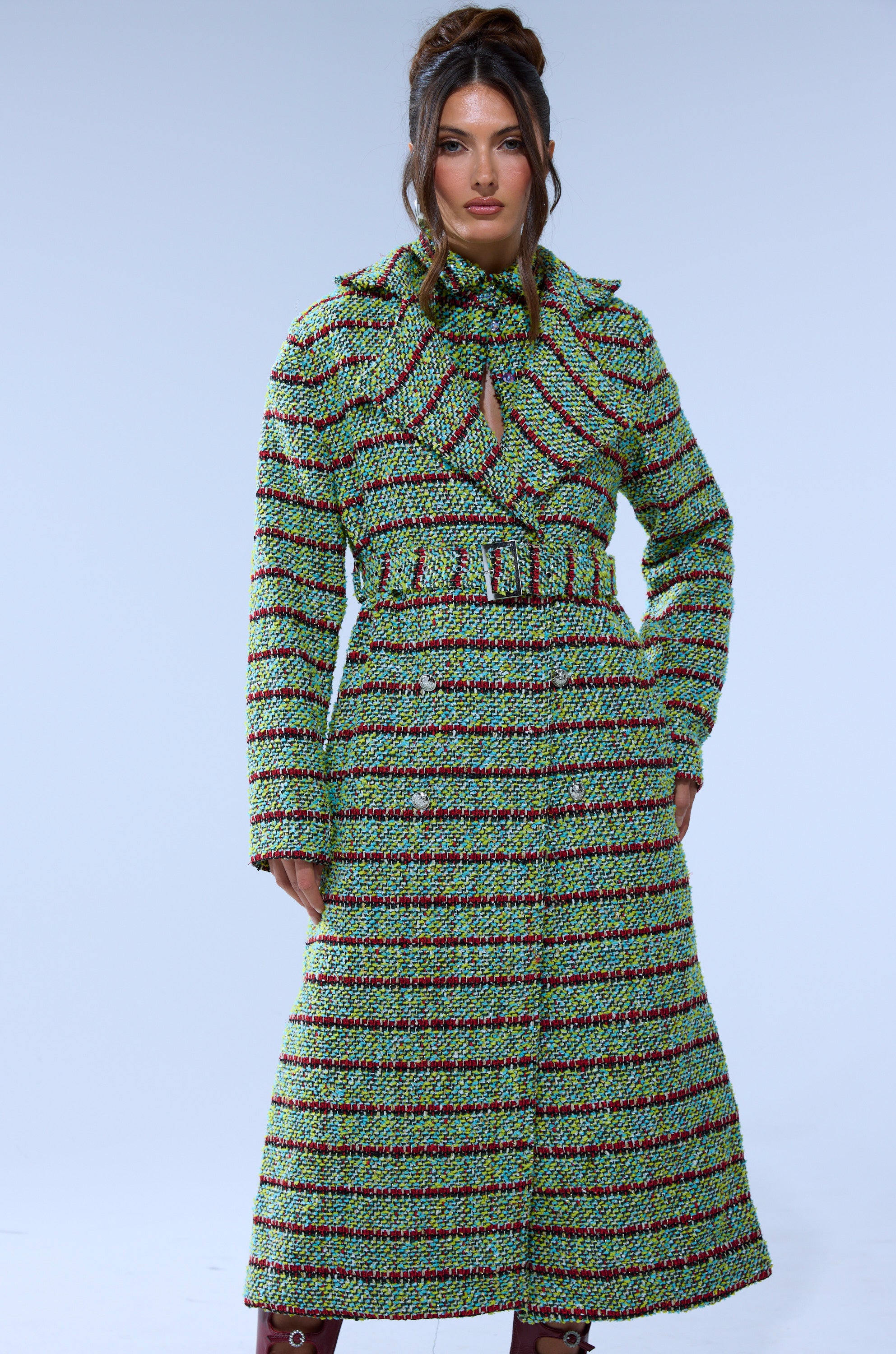 A woman poses against a plain background in the IT'S GIVING LUCK TWEED TRENCH, a green and black striped coat with a belt and wide collar. Her long, wavy hair is styled in a partial updo as she looks directly at the camera.