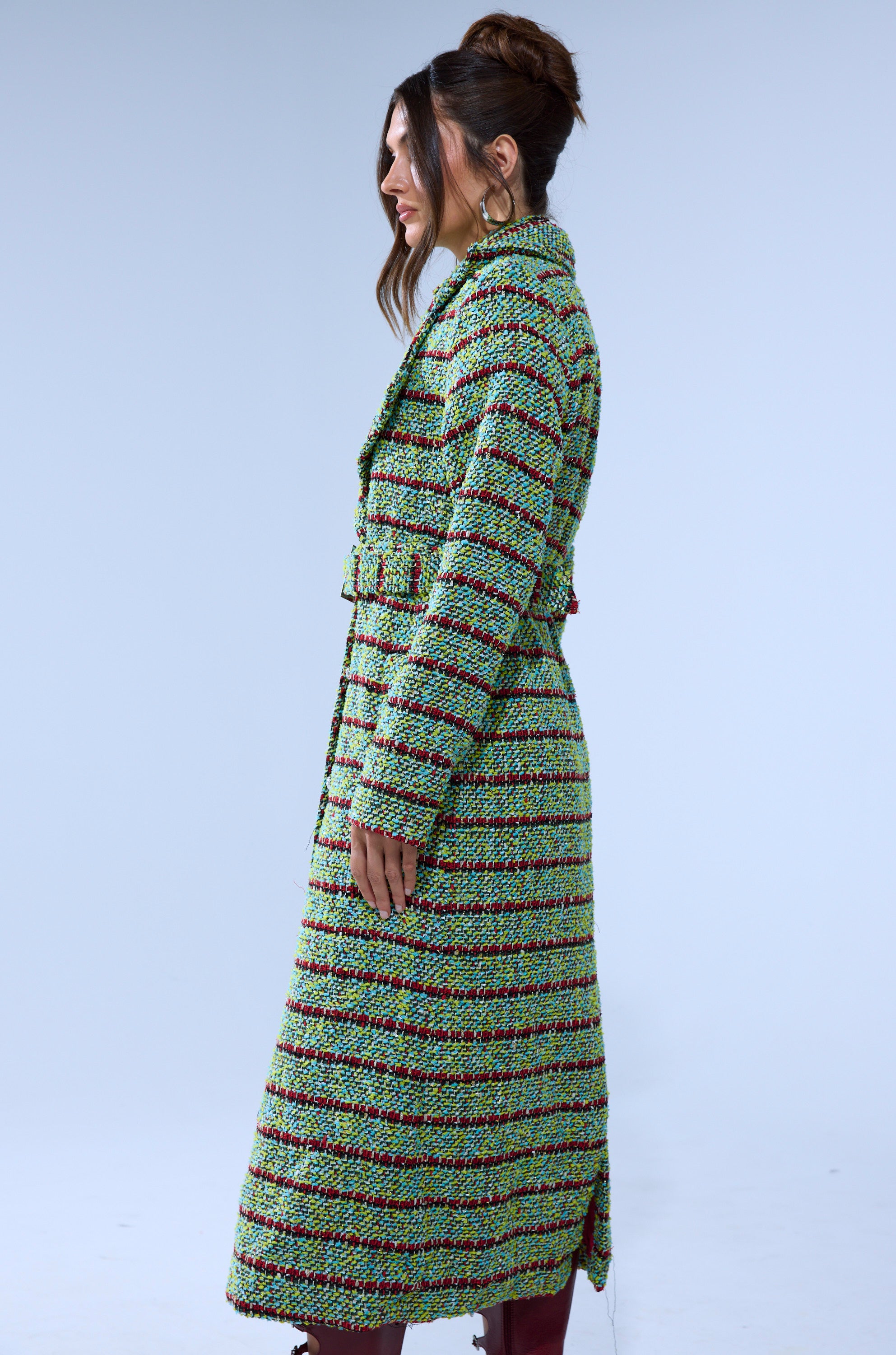 A woman stands in profile against a plain background, wearing the IT'S GIVING LUCK TWEED TRENCH with red and black stripes, her hair in a loose bun, styled with maroon boots.
