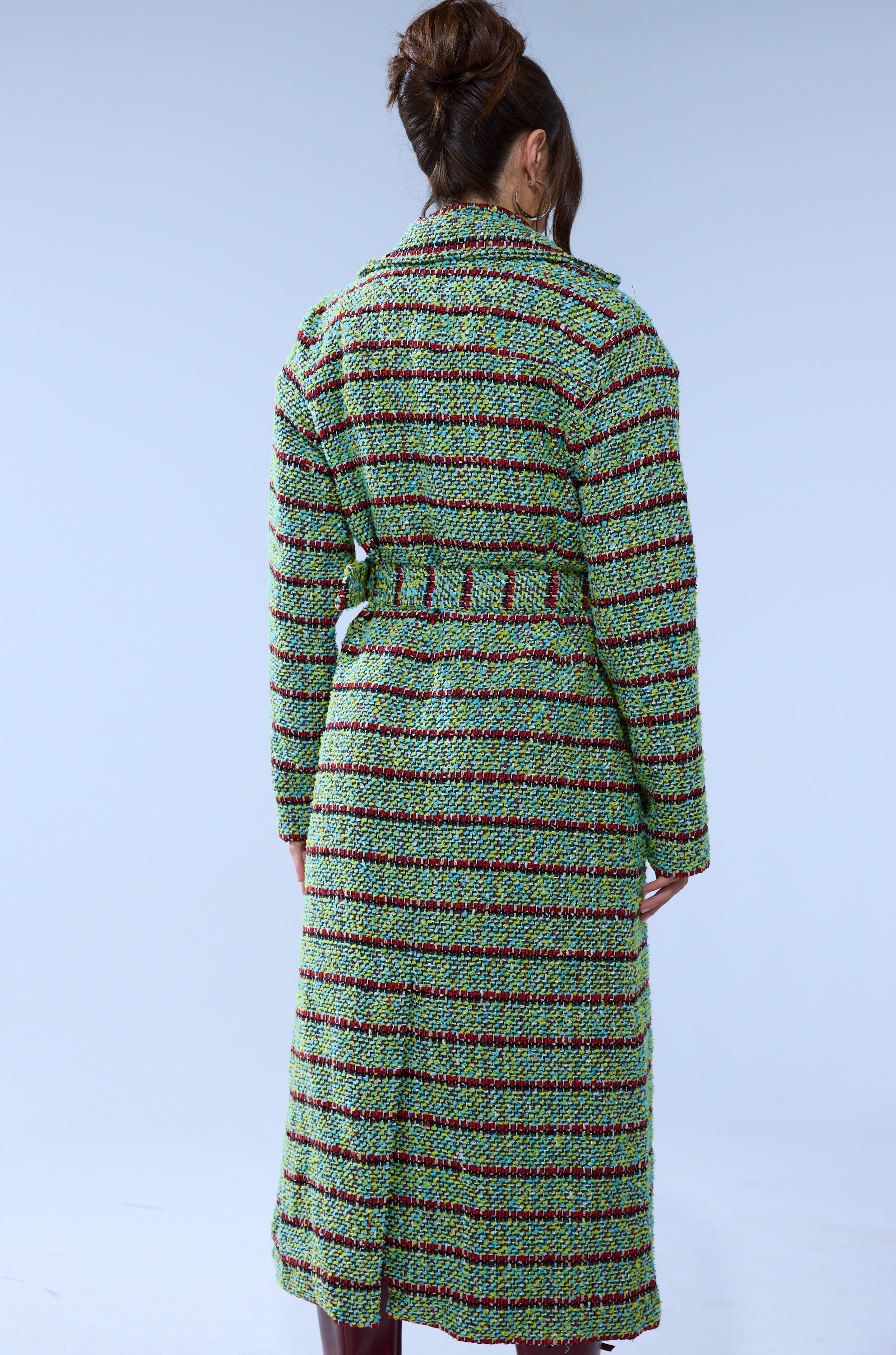 A woman with brown hair in a bun is seen from the back, wearing the IT'S GIVING LUCK TWEED TRENCH, a multicolored plaid belted coat, standing against a plain light blue background.