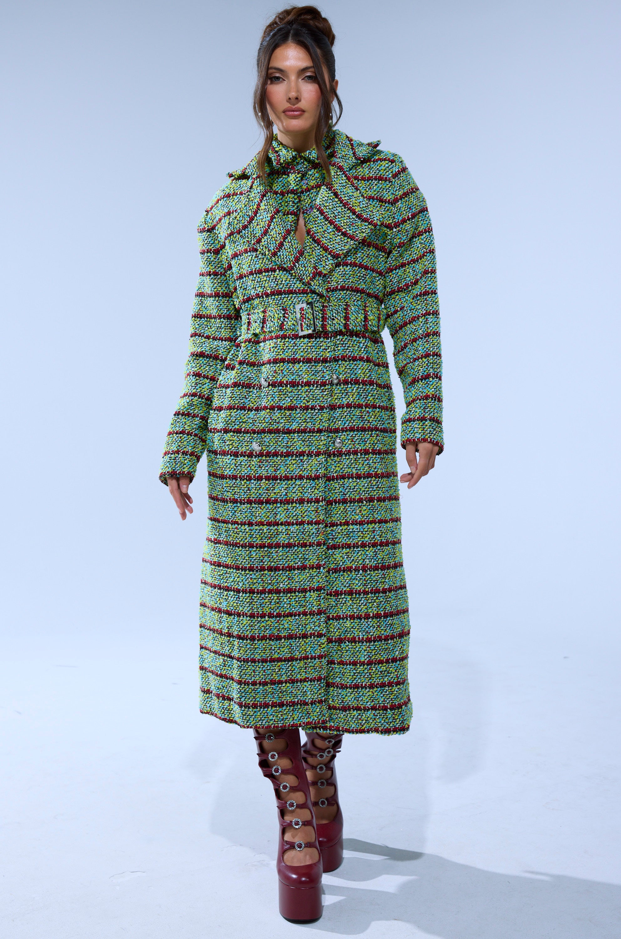 A woman wears the IT'S GIVING LUCK TWEED TRENCH—a longline green and pink patterned coat with a matching scarf—paired with brown platform boots with circular cutouts, walking against a plain, light-colored background.