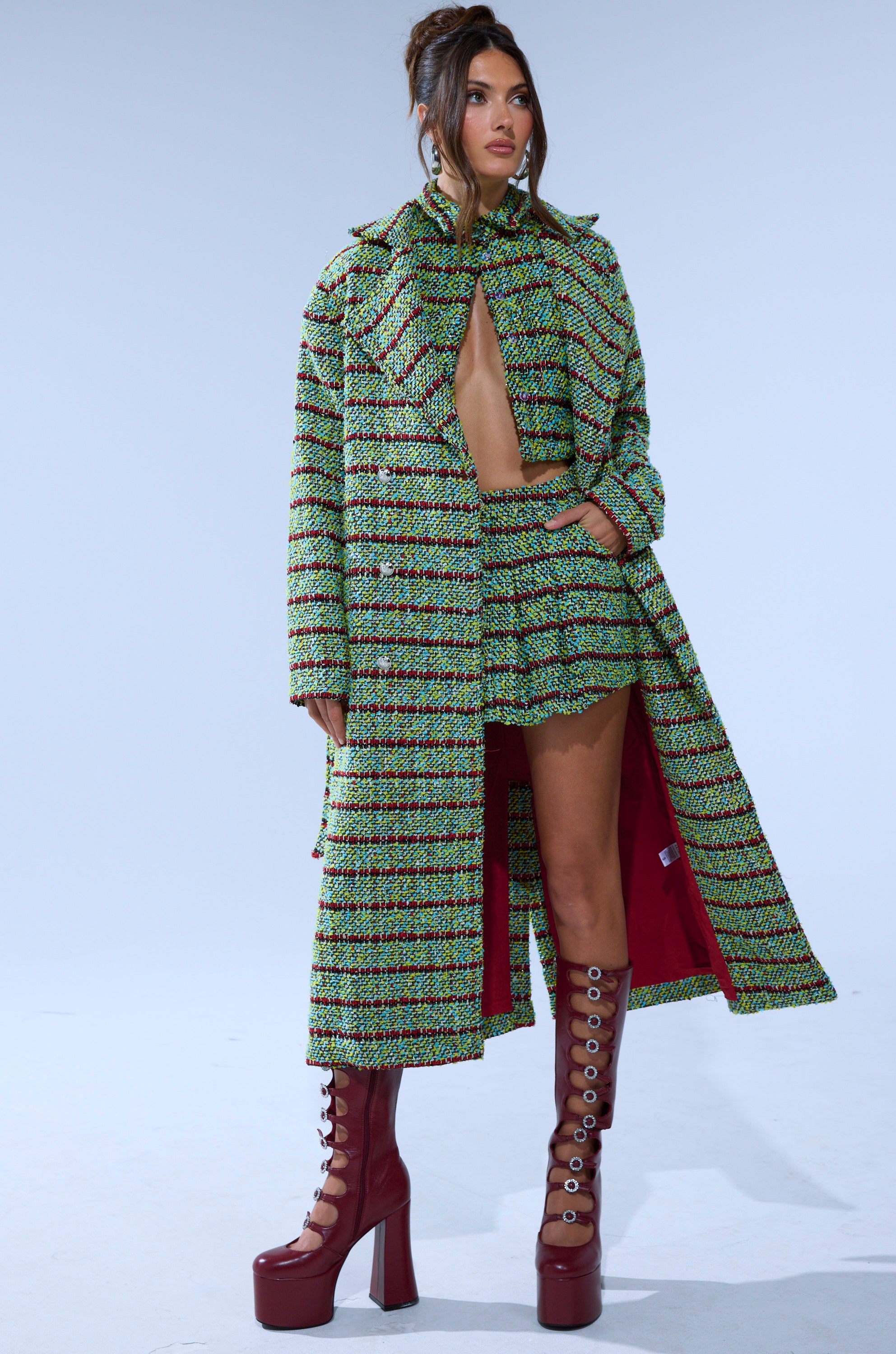 A woman exudes city-chic confidence in the IT'S GIVING LUCK TWEED TRENCH, a green, yellow, and black patterned coat paired with a matching cropped top, mini skirt, and tall maroon platform boots against a simple backdrop.