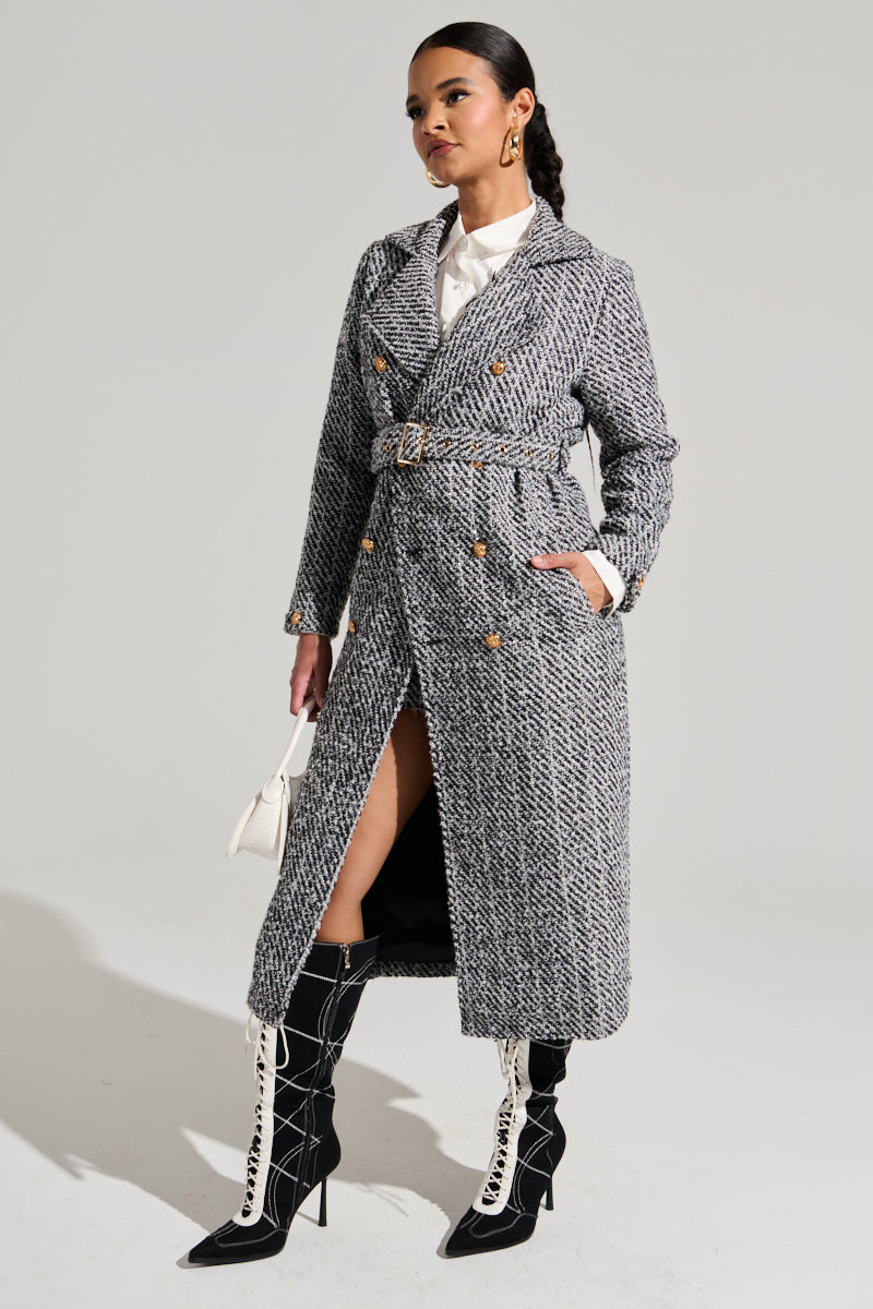 A woman stands confidently against a plain background, wearing the ALTURA TWEED TRENCH from AKIRA Label, featuring double-breasted front button closures and a waist belt over a white shirt. She pairs it with black knee-high, lace-up boots with high heels and holds a small white handbag.