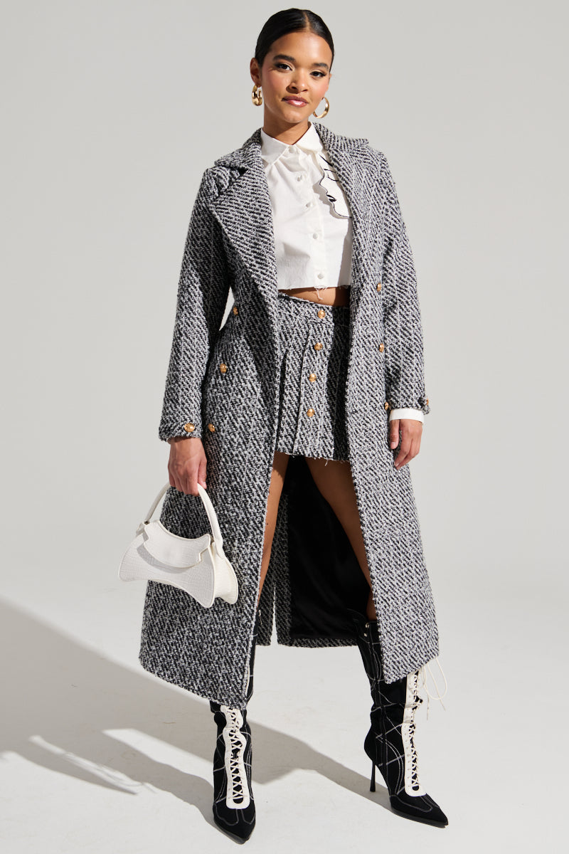 A woman stands confidently posing, wearing the ALTURA TWEED TRENCH from AKIRA Label, featuring a tweed waist belt over a white blouse and matching skirt. Her knee-high black lace-up boots complement her small white handbag. With neatly styled hair and elegant earrings, she exudes sophistication.