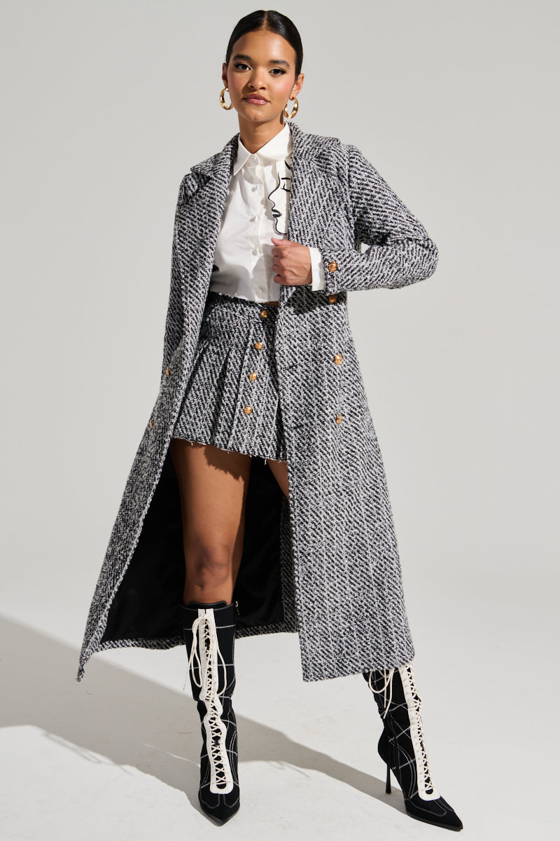A person poses confidently in a stylish outfit consisting of the AKIRA Label ALTURA TWEED TRENCH with double-breasted front button closures and a tweed waist belt, paired with a white collared shirt, a short pleated skirt, and knee-high lace-up boots. They complement their ensemble with hoop earrings and have their hair pulled back.
