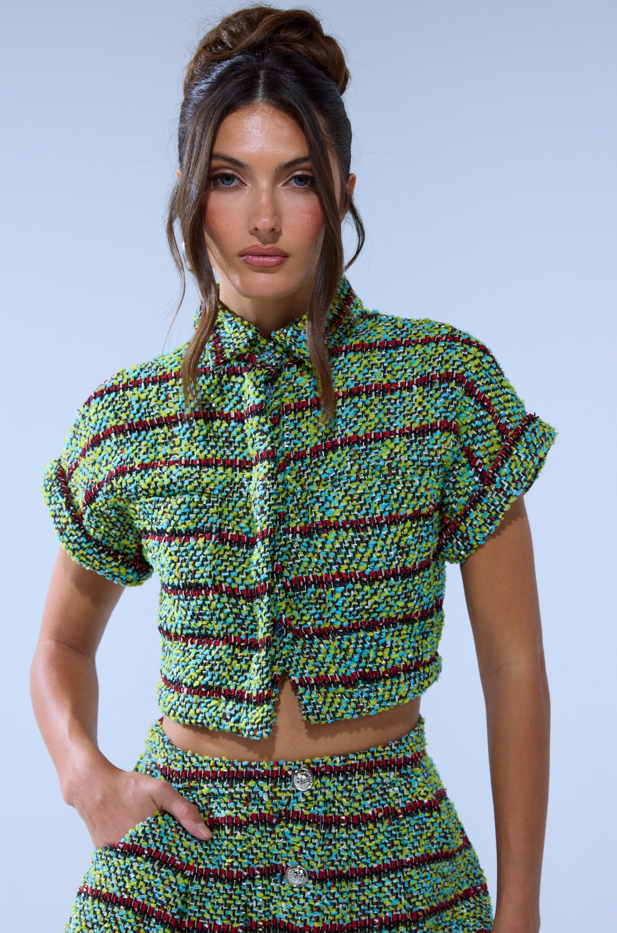 A woman with long wavy brown hair styled in an updo wears the IT'S GIVING TWEED BUTTON DOWN and matching high-waisted shorts in green, blue, and black plaid, standing against a plain light blue background.