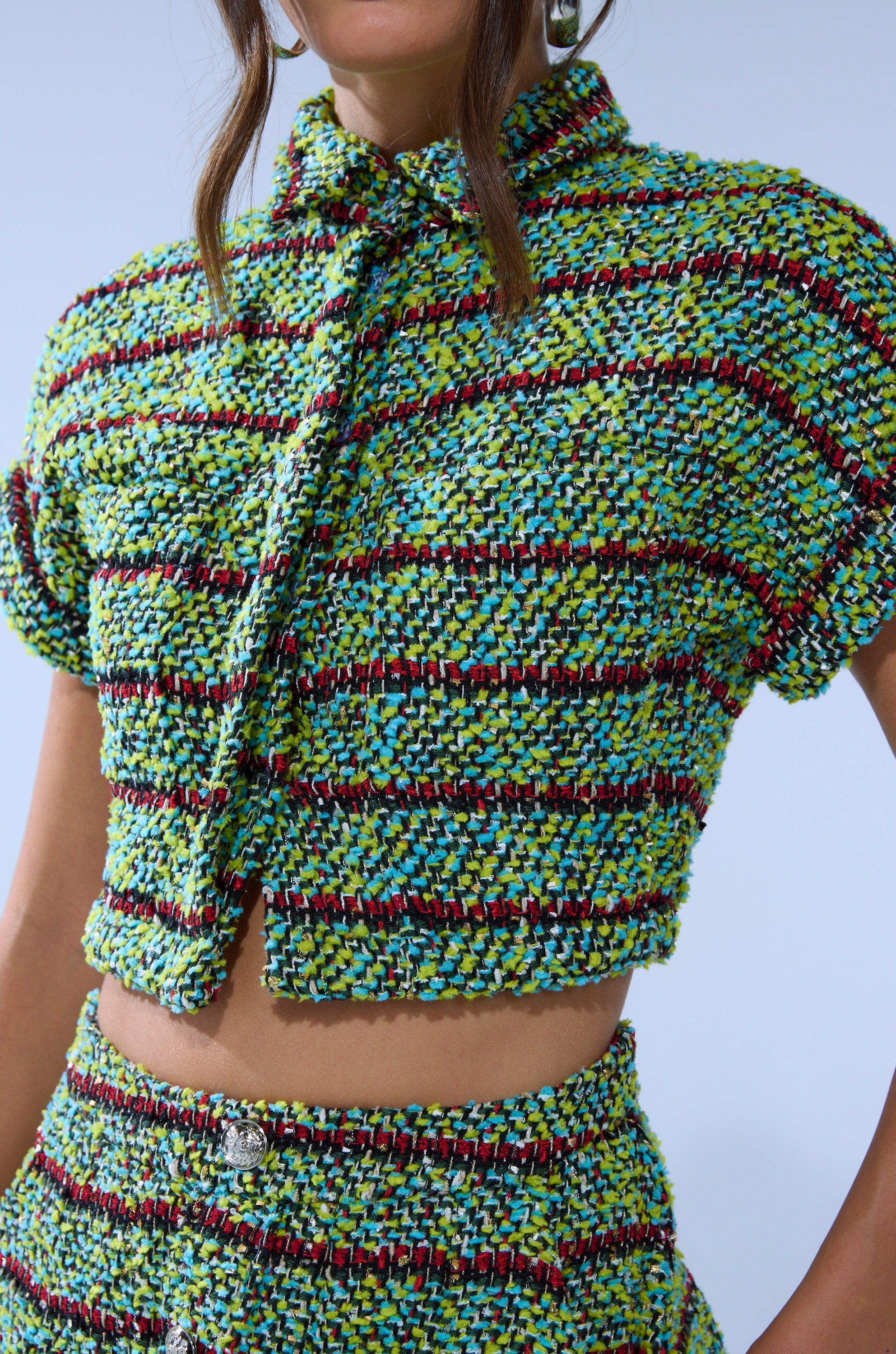 A woman wears the IT'S GIVING TWEED BUTTON DOWN, a colorful cropped tweed shirt with short sleeves and collar, paired with a matching high-waisted skirt in vibrant green, blue, yellow, red, and black plaid.
