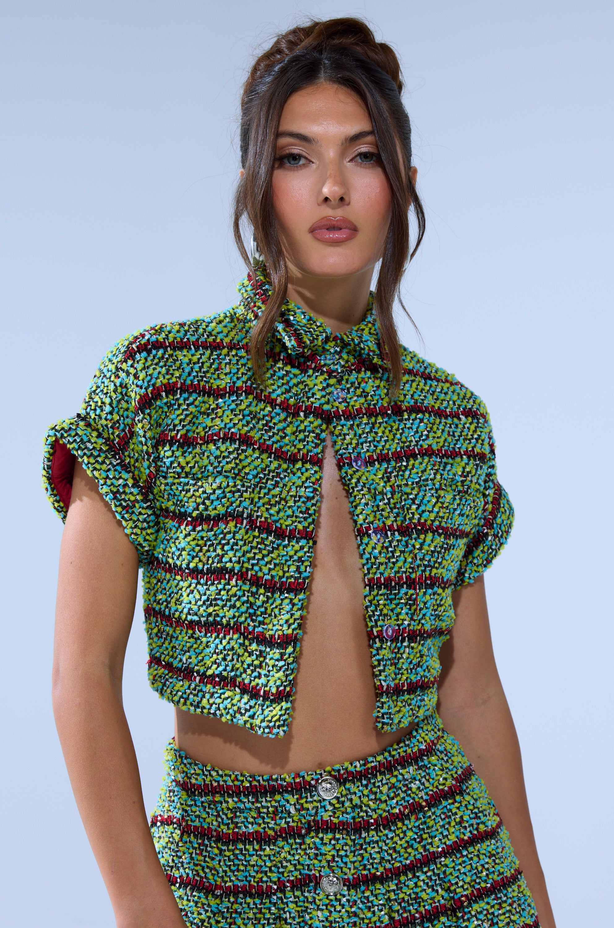 A woman with long dark wavy hair wears the IT'S GIVING TWEED BUTTON DOWN cropped jacket and matching skirt in green, blue, and maroon with metallic buttons, posing against a light blue background.