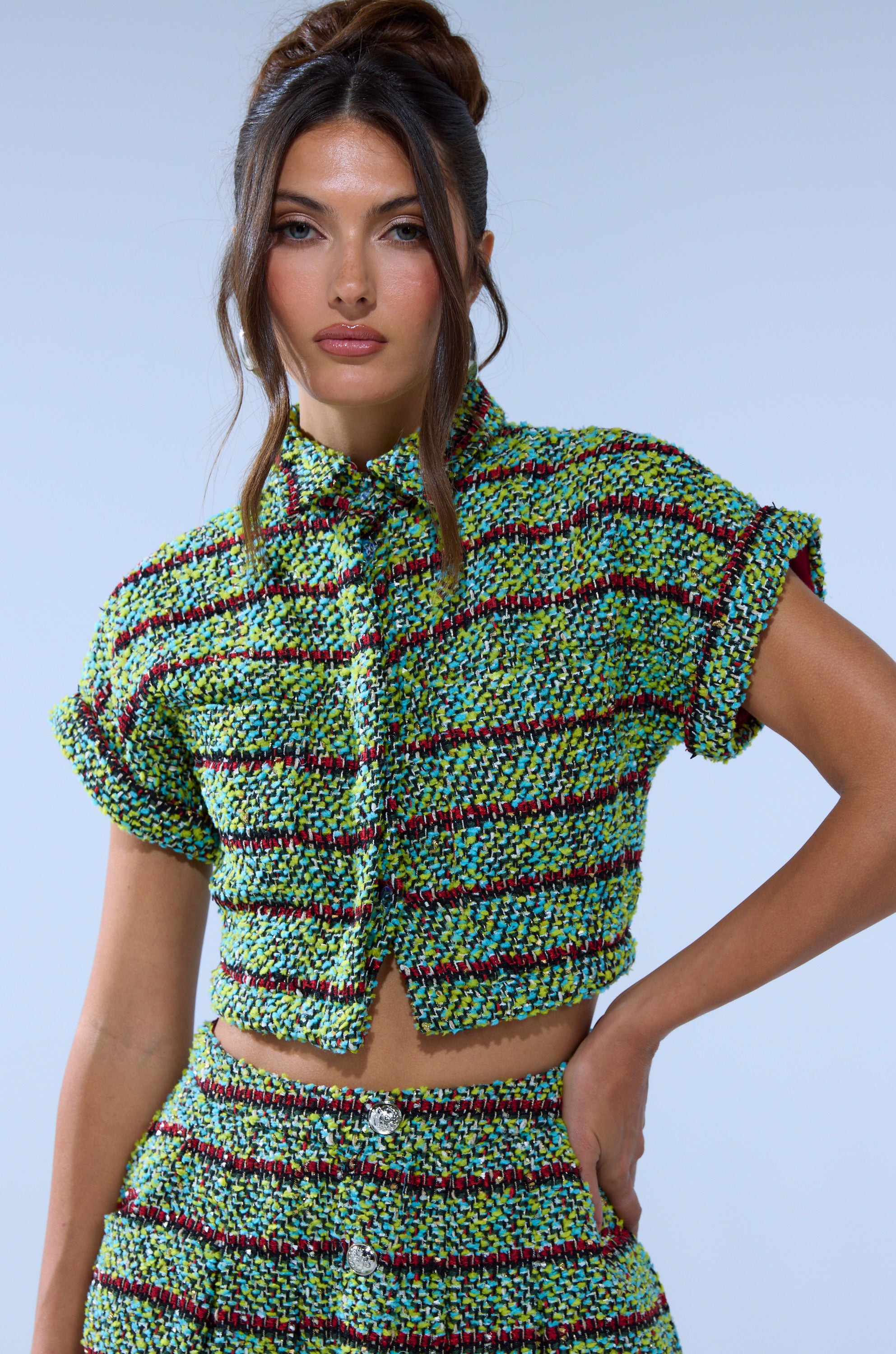 A woman with long brown hair in a loose updo poses with one hand on her hip against a light blue background, wearing the IT'S GIVING TWEED BUTTON DOWN plaid cropped shirt and matching skirt in green, yellow, black, and blue stripes.