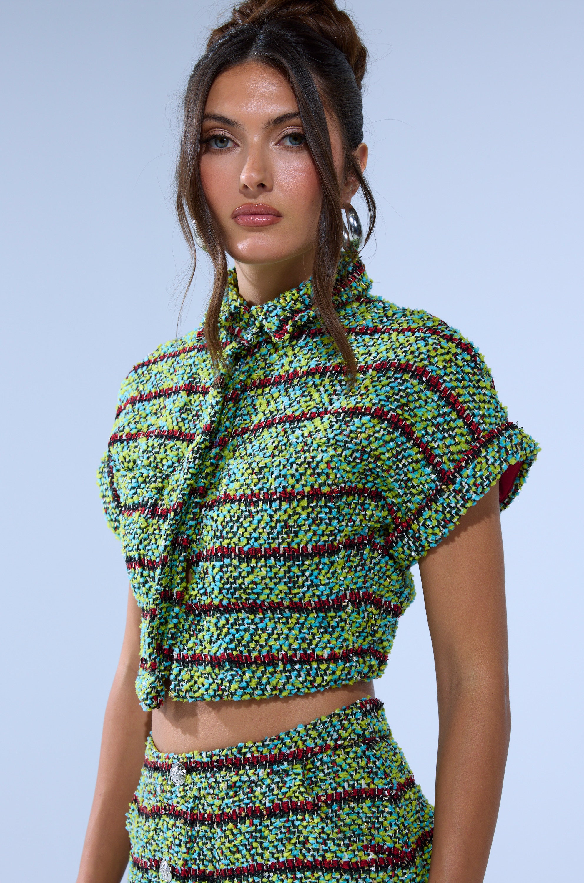 A woman with long brown hair in a loose updo wears the IT'S GIVING TWEED BUTTON DOWN, a cropped shirt in textured, multicolored fabric with green, yellow, and purple stripes, paired with a matching high-waisted skirt.
